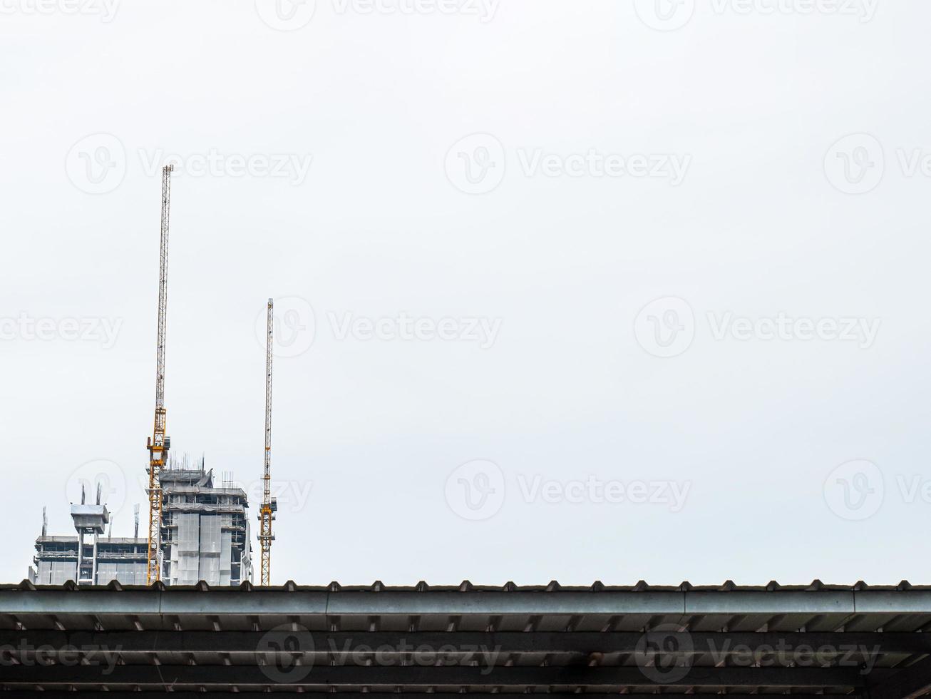 Two construction cranes are working on the construction of tall buildings, lifting and placing things. The construction site is covered with blankets. in the white sky city building in a straight line photo