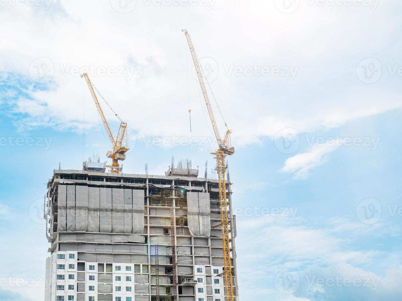 Two construction cranes are working on the construction of tall buildings, lifting and placing things. The construction site is covered with blankets. in the clear sky city building in a straight line photo