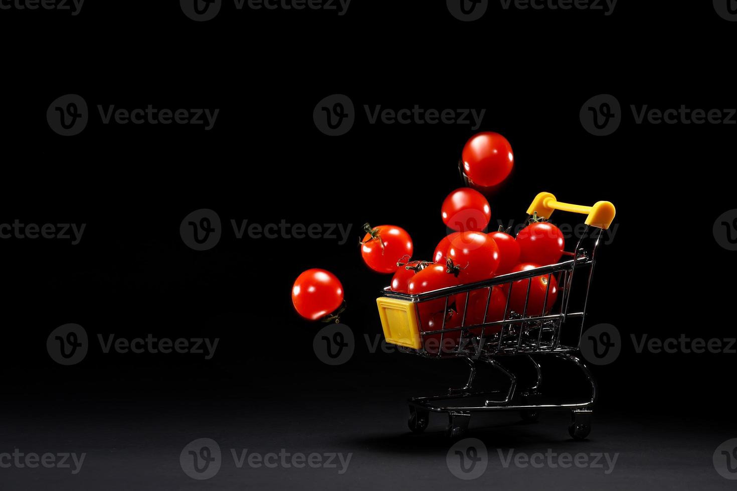 Mini shopping cart with fresh cherry tomatoes on black background. Supermarket advertisement copy space. Grocery shop. Tomatoes delivery. Transporting goods. Retail and selling. photo
