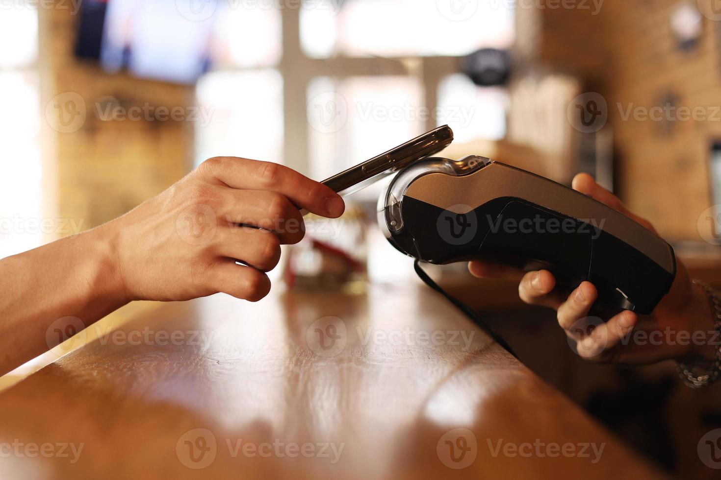 Macro unknown hand giving terminal to visitor for mobile phone payment indoor. Client hand paying bill with smartphone in blurred background. Cropped shot hands making contactless transaction inside. photo