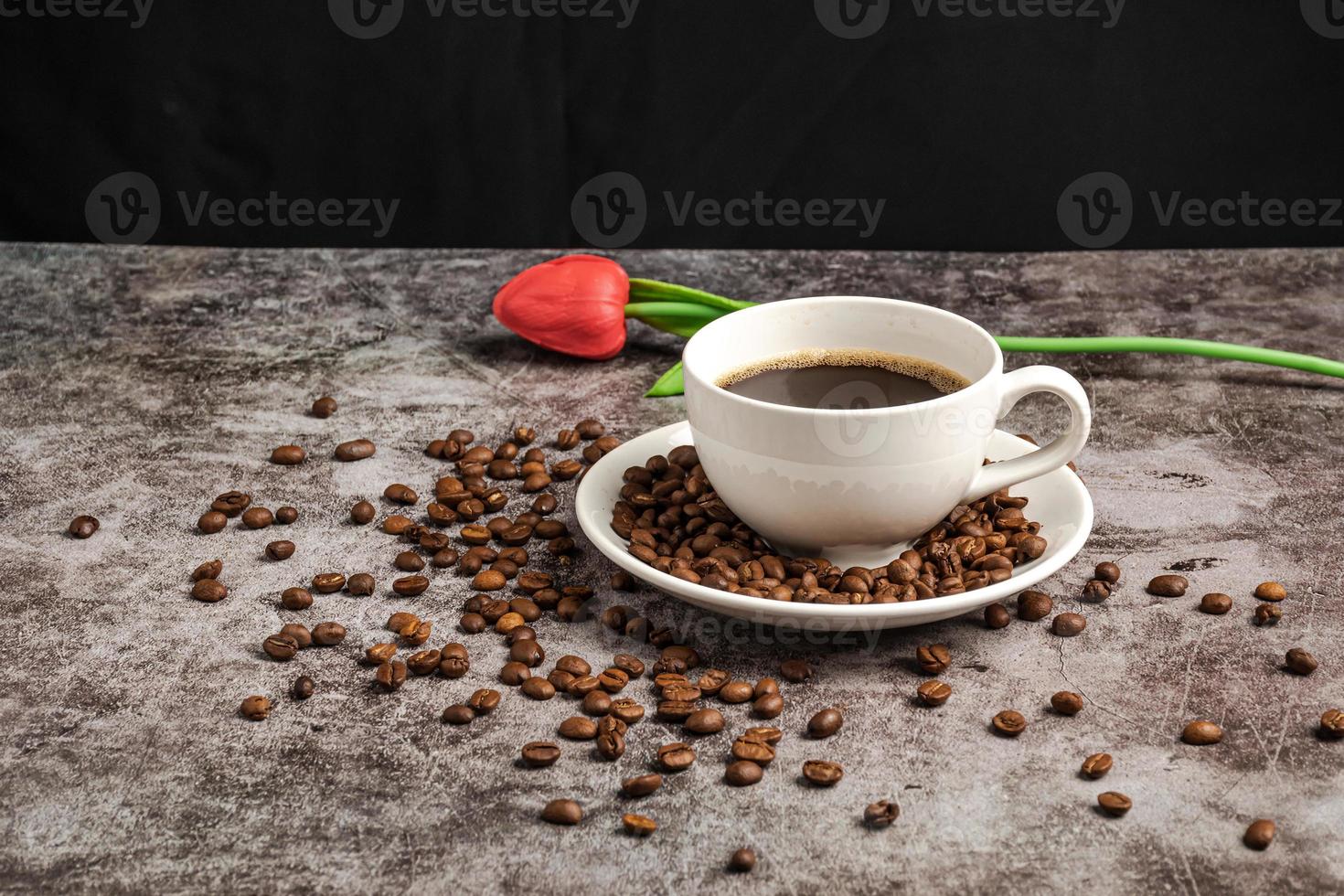 A close-up of hot coffee in a white cup is placed on a cement floor table, lots of roasted coffee beans are in the coffee cup saucer, and around, smoke and aroma waft from the cup. Blurred background photo