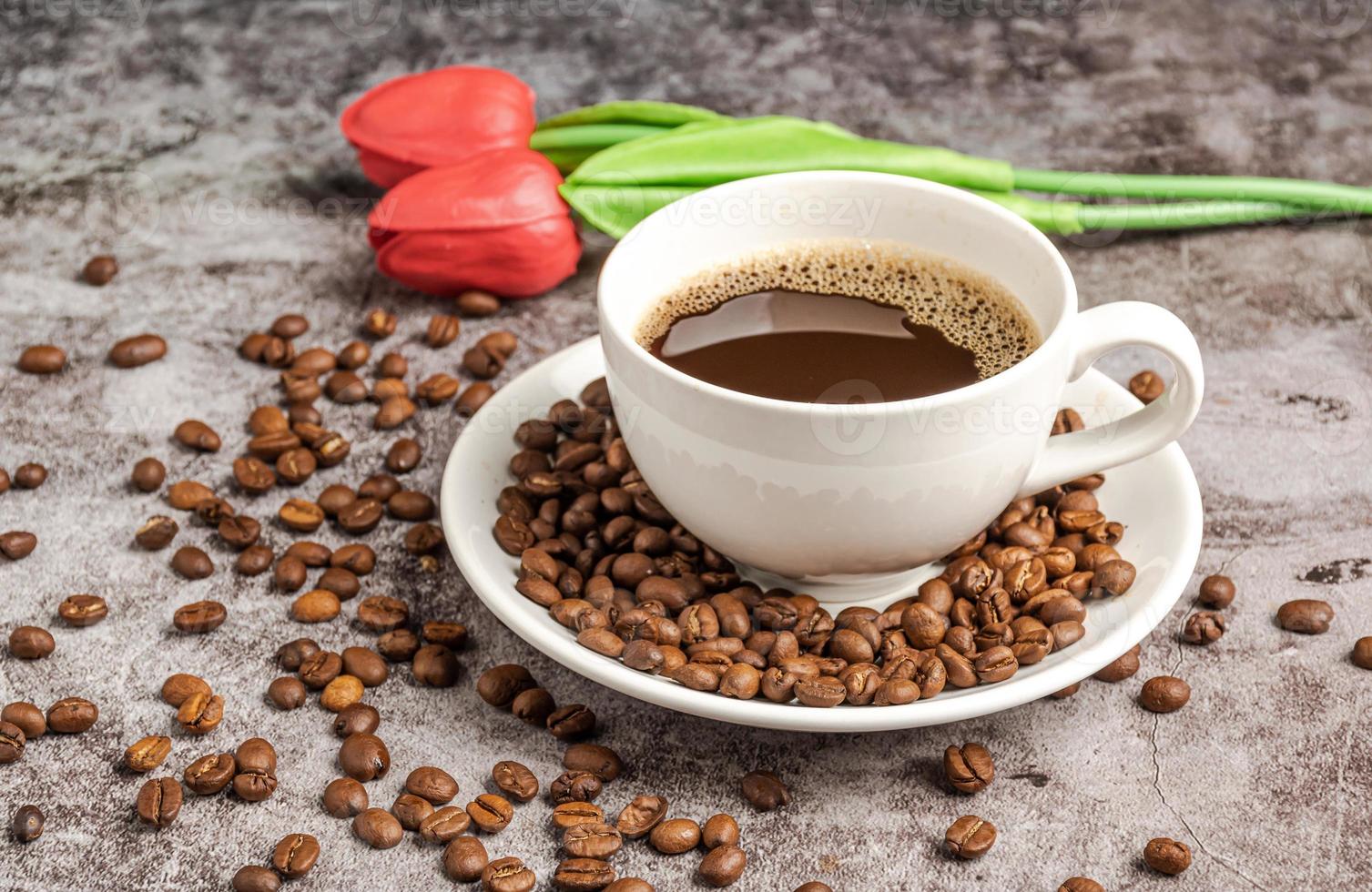 A close-up of hot coffee in a white cup is placed on a cement floor table, lots of roasted coffee beans are in the coffee cup saucer, and around, smoke and aroma waft from the cup. Blurred background photo