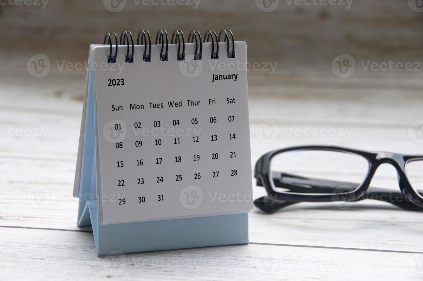 enero de 2023 calendario de escritorio blanco con gafas sobre fondo de mesa de madera. copie el espacio foto