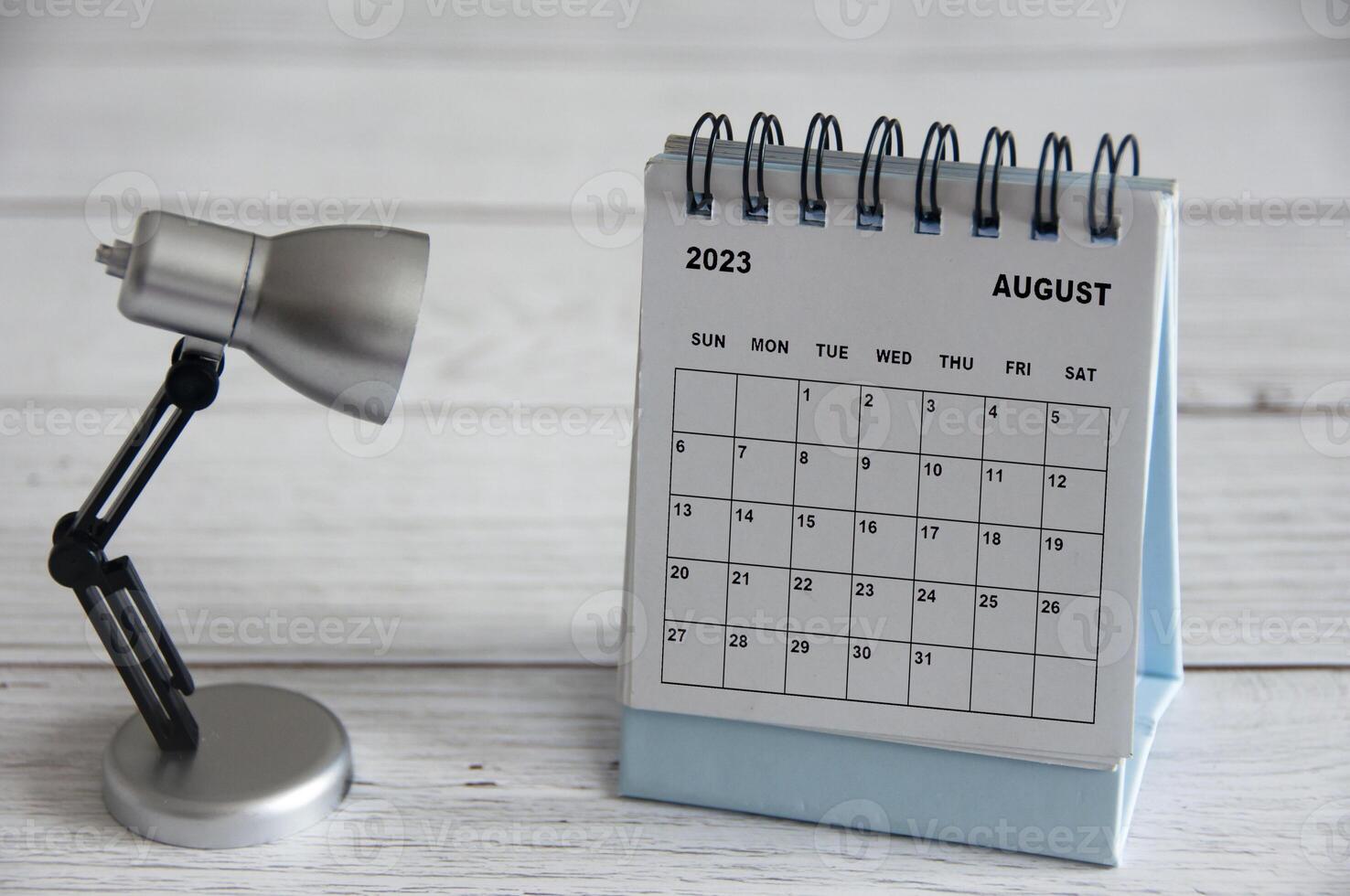 agosto de 2023 calendario de escritorio blanco con lámpara de mesa sobre mesa de madera. foto