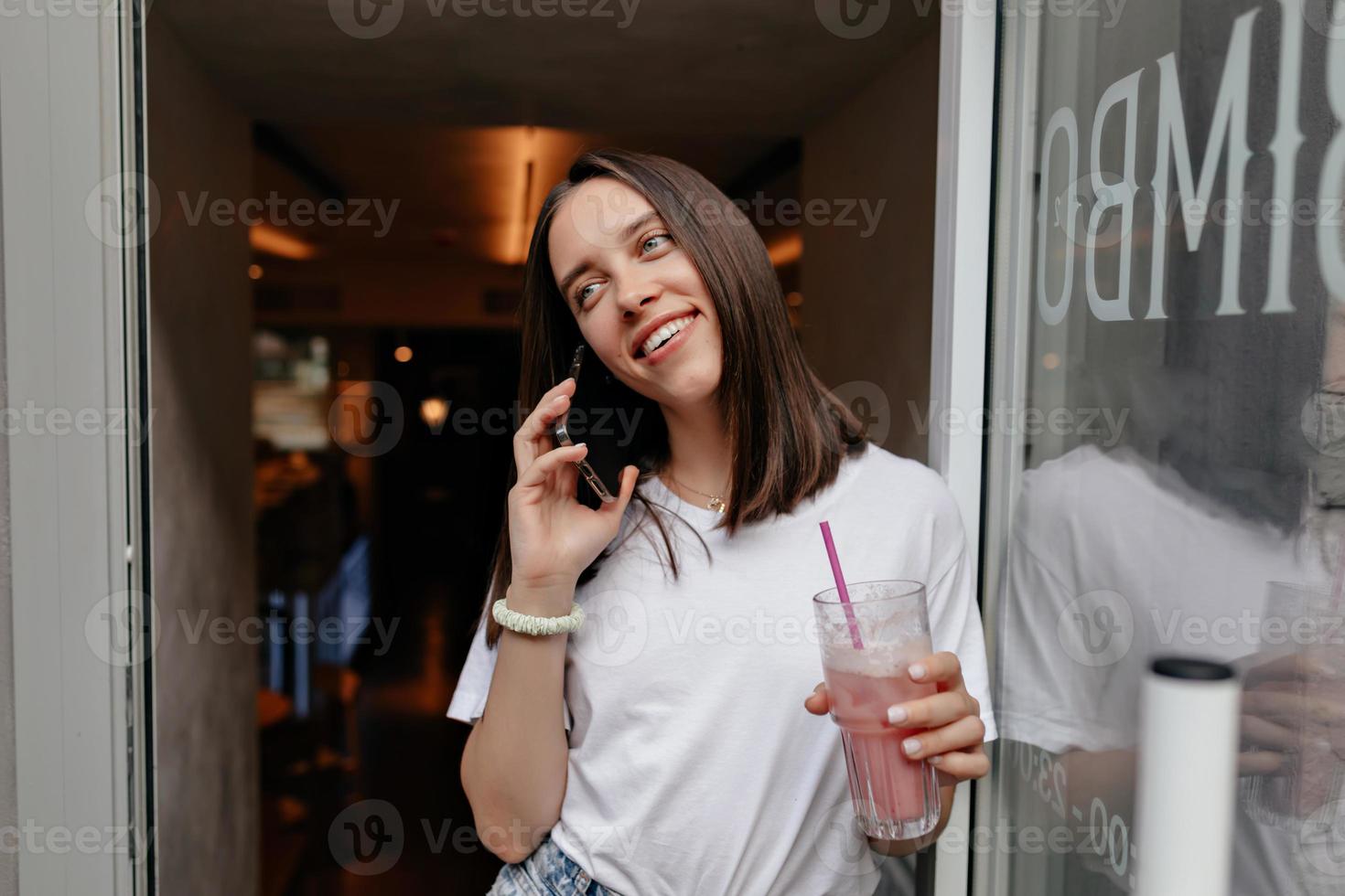 Adorable happy girl with wonderful smile is talking on the phone and drinking bright summer smoothie while come out from cafeteria photo