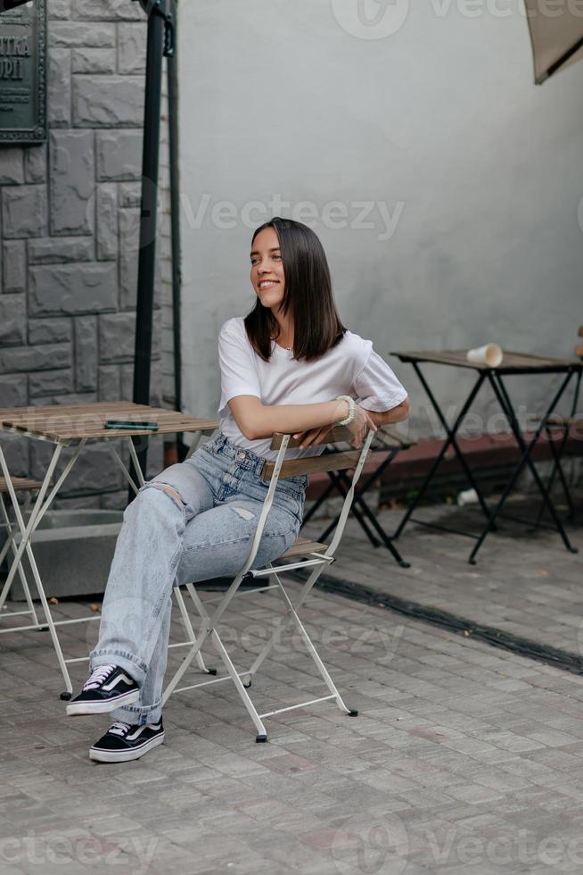 Portrait of pretty young caucasian woman warming up on autumn day. Brunette in white t-shirt, jeans and sports shoes sits in cafe. Rest and recovery concept. photo