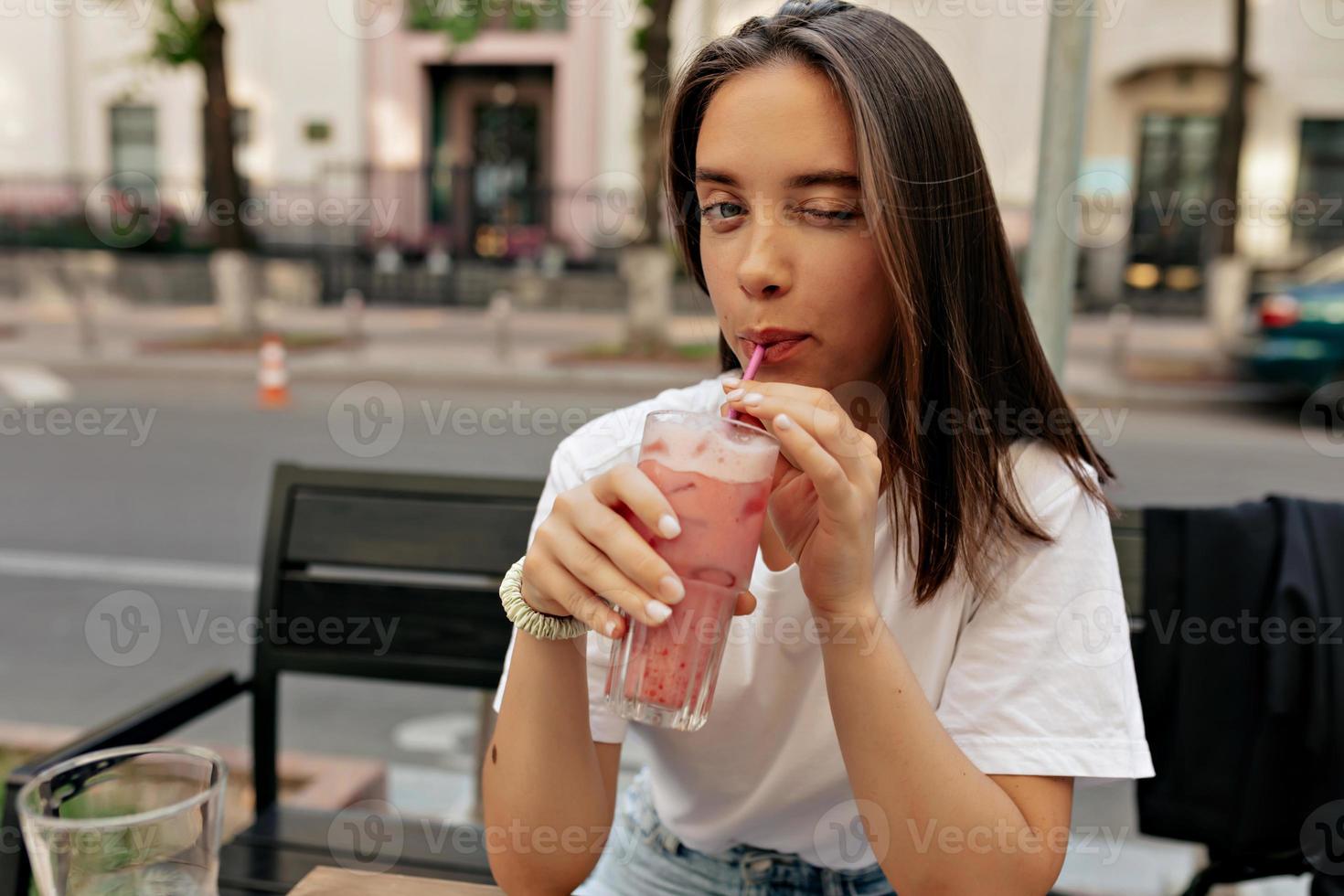 seductora mujer joven guiñando un ojo a la cámara y sonriendo mientras bebe un batido de verano y se sienta en el primer plano de la terraza. las curvas desnudas y delgadas de las clavículas son llamativas. foto