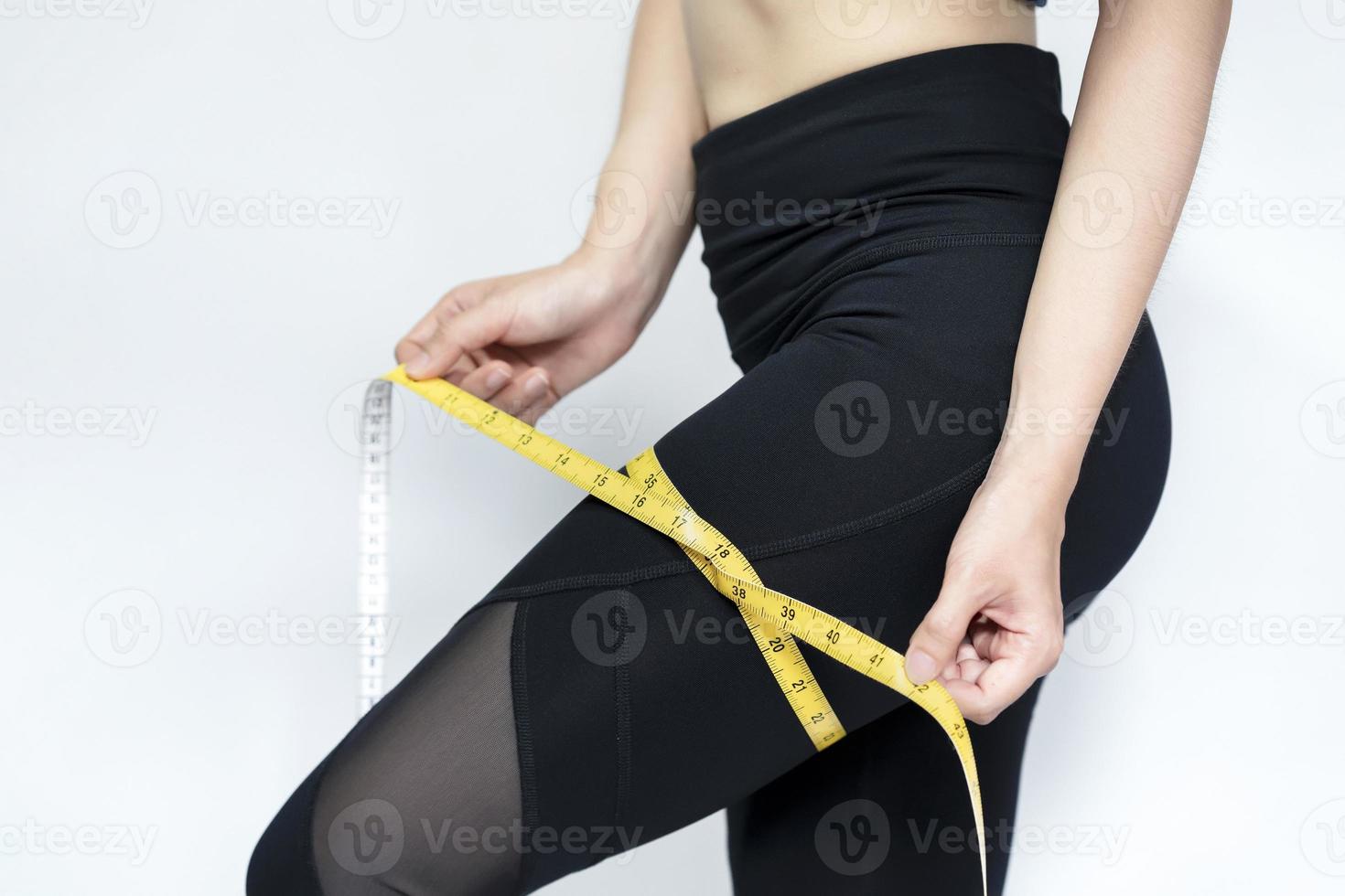 A skinny woman wearing black pants uses a tape measure around her thigh against with gray background. Health care concept. photo