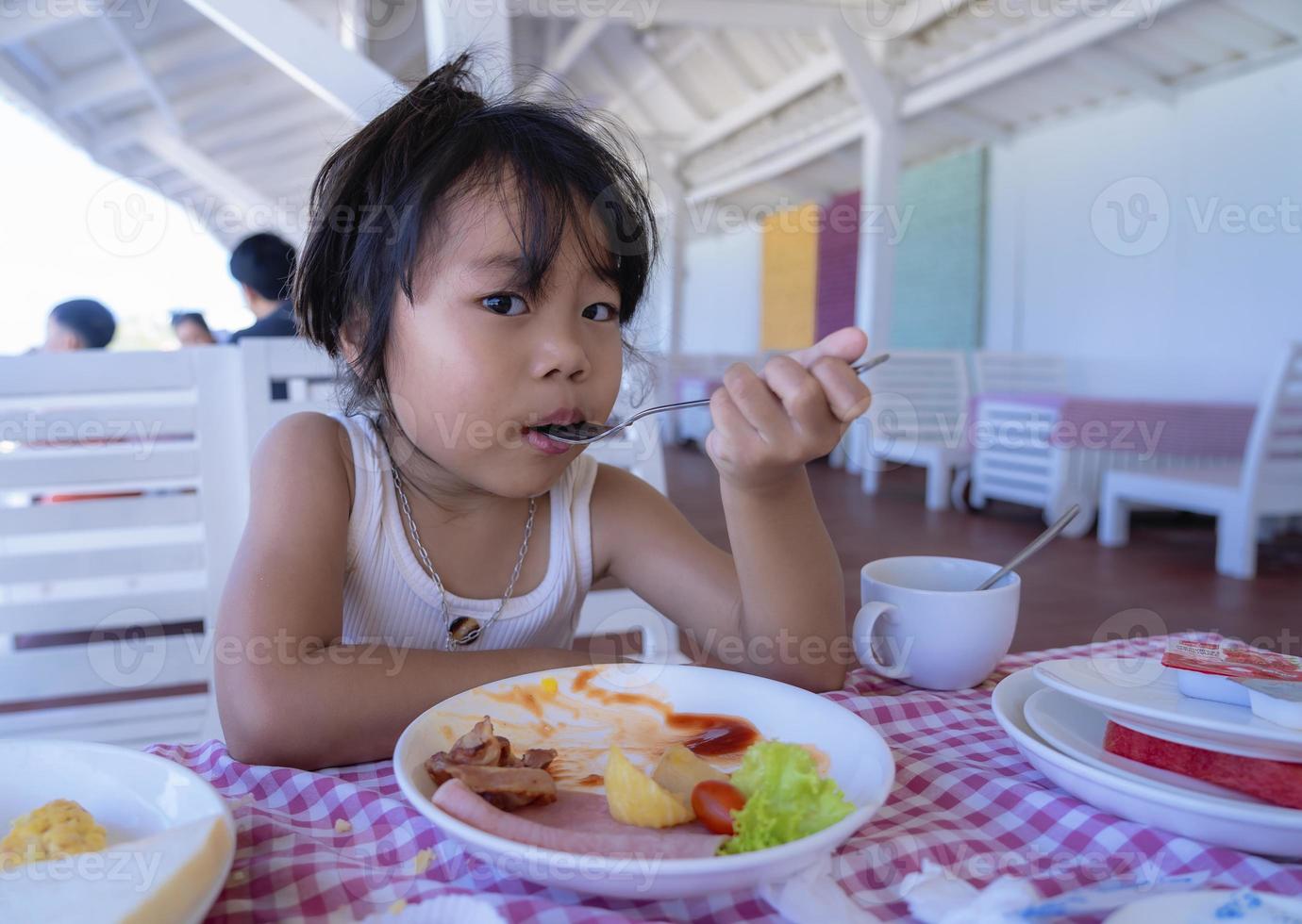 Cute little child girl eating breakfast holding spoon and stains food around her mouth and looking at camera. Hungry little woman breakfast time at restaurant. Kid and Healthy Eating concept. photo