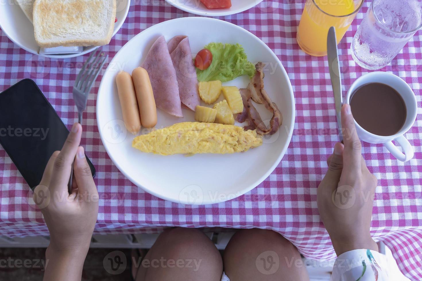 mesa de desayuno fresca con rollo de tortilla, salchicha, tocino, jamón, tomate y patata en un plato. las manos de una mujer que sostenían un tenedor y un cuchillo incluían café, jugo de naranja, pan y teléfono móvil en una mesa. foto