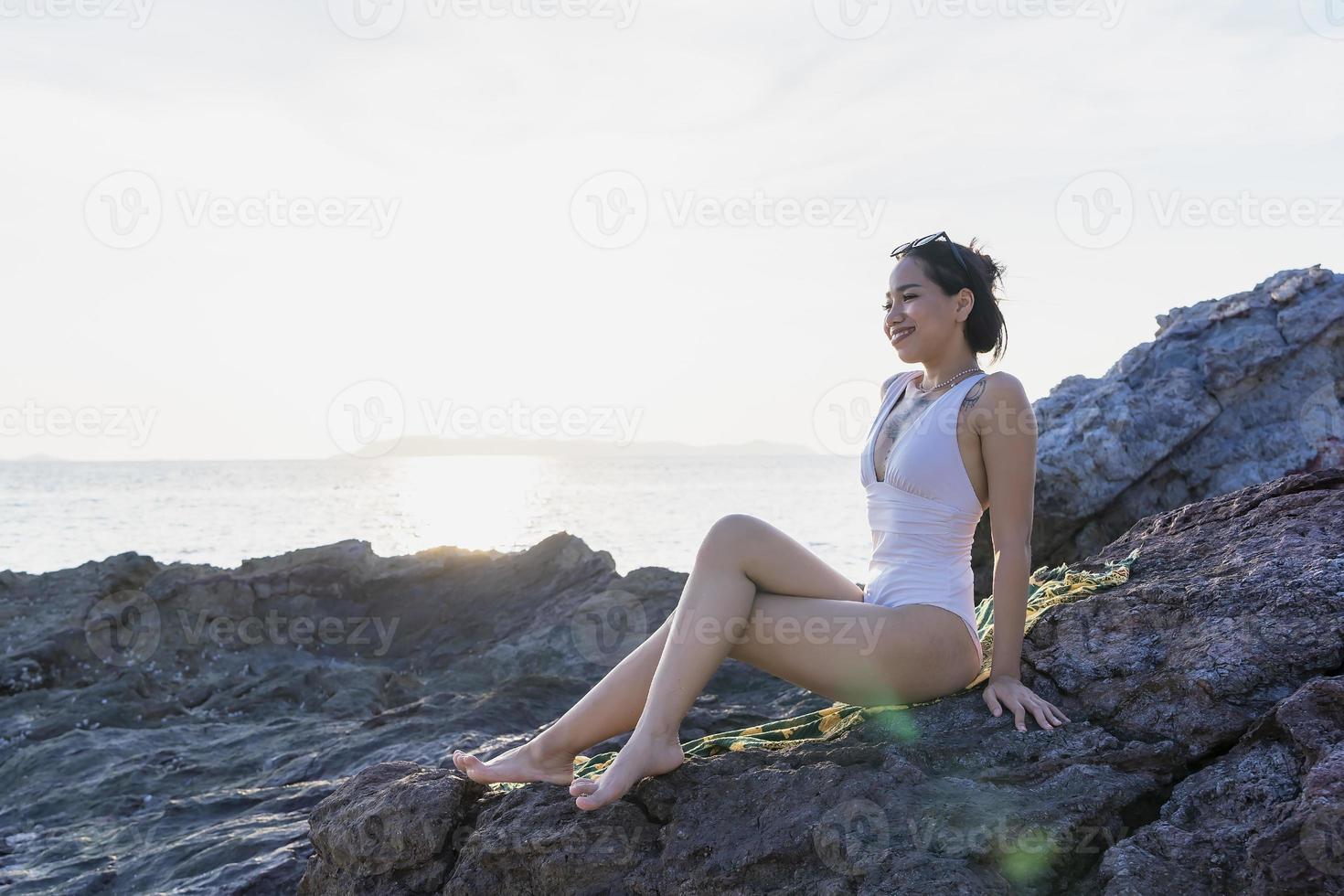 Portrait of beautiful fashionable woman in white bikini posing at the rock beach. Young Asian woman dressed in swimsuit laying on a rock on a summer day with sunset backgrounds at Koh Larn in Pattaya photo