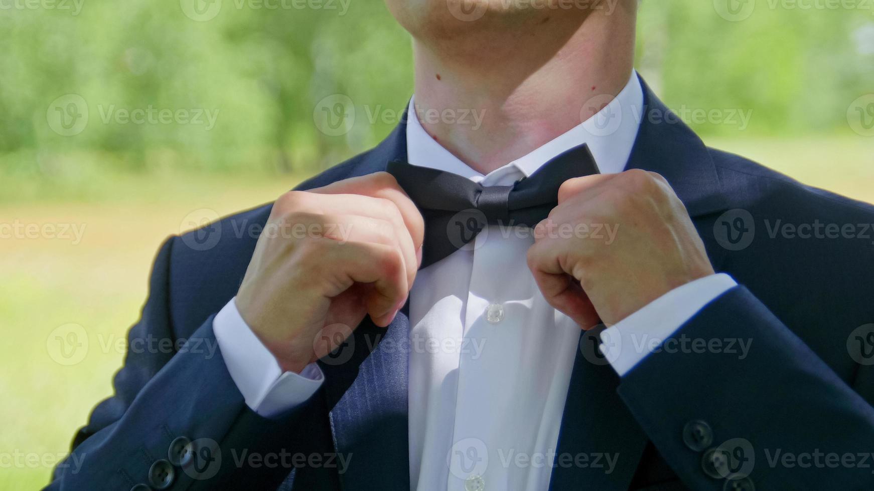 The groom in a white shirt puts on bow tie from the front, outdoor, closeup. photo