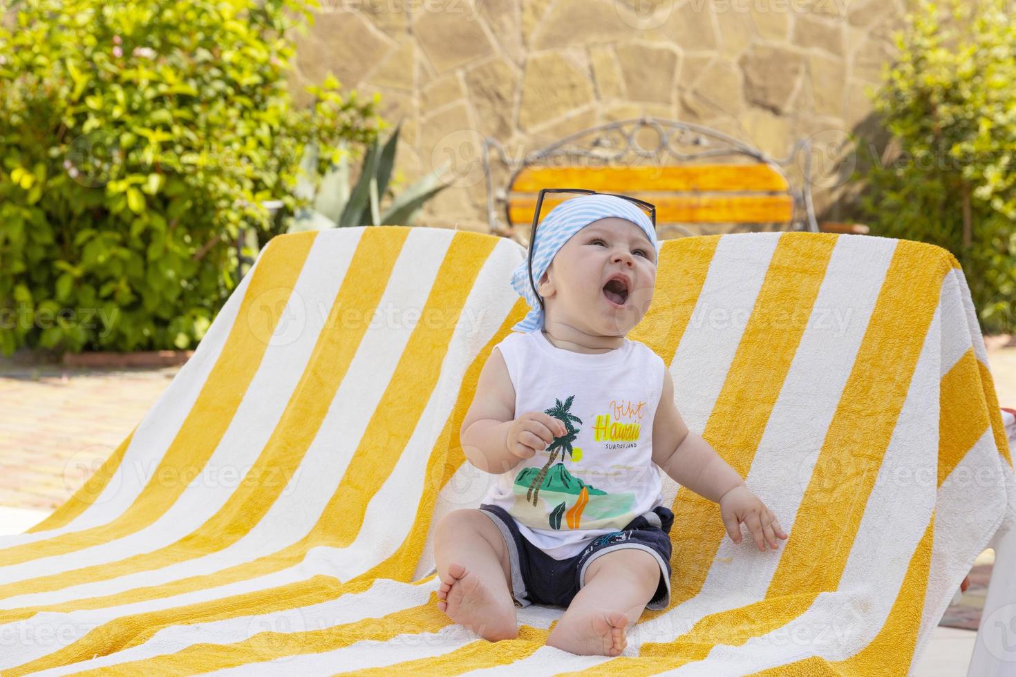 Handsome little boy in sunglasses and a bandana sits on a sun lounger by the pool. Selective focus. photo