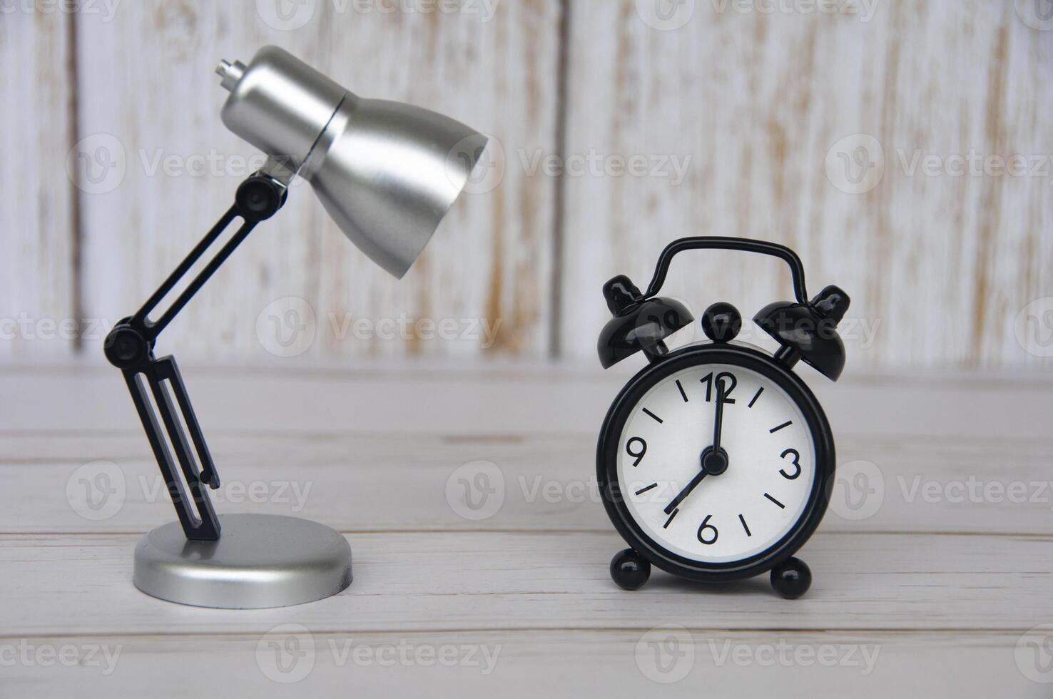 Alarm clock pointing at 7 o'clock with table lamp on wooden background. photo