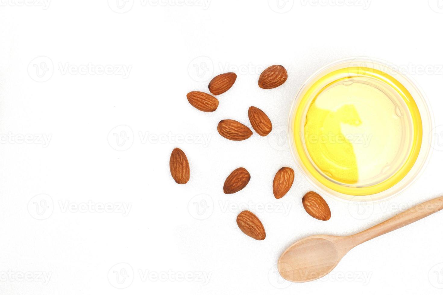 Clear yellow almond oil in a clear bowl with a wooden spoon on top of it and several almonds on a white background.top view, flat lay, top-down. photo