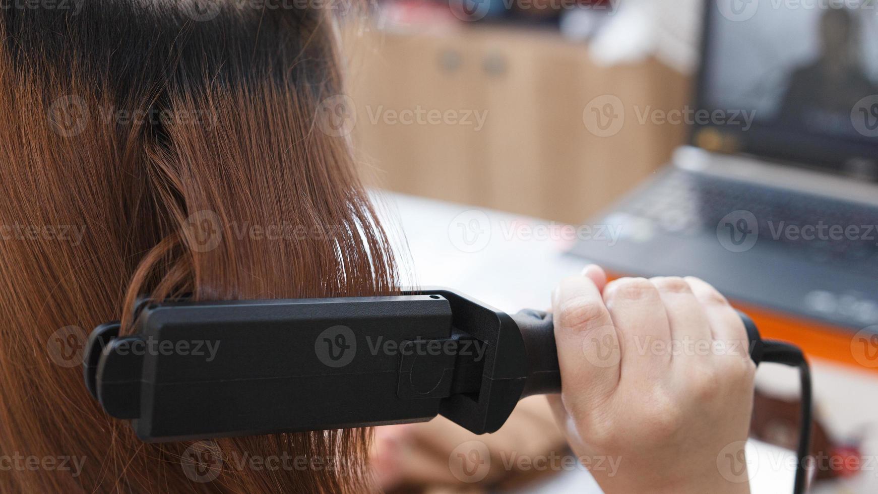 detrás de escena de una mujer de pelo rojizo que se alisa el pelo con una plancha en la cama de su casa. foto