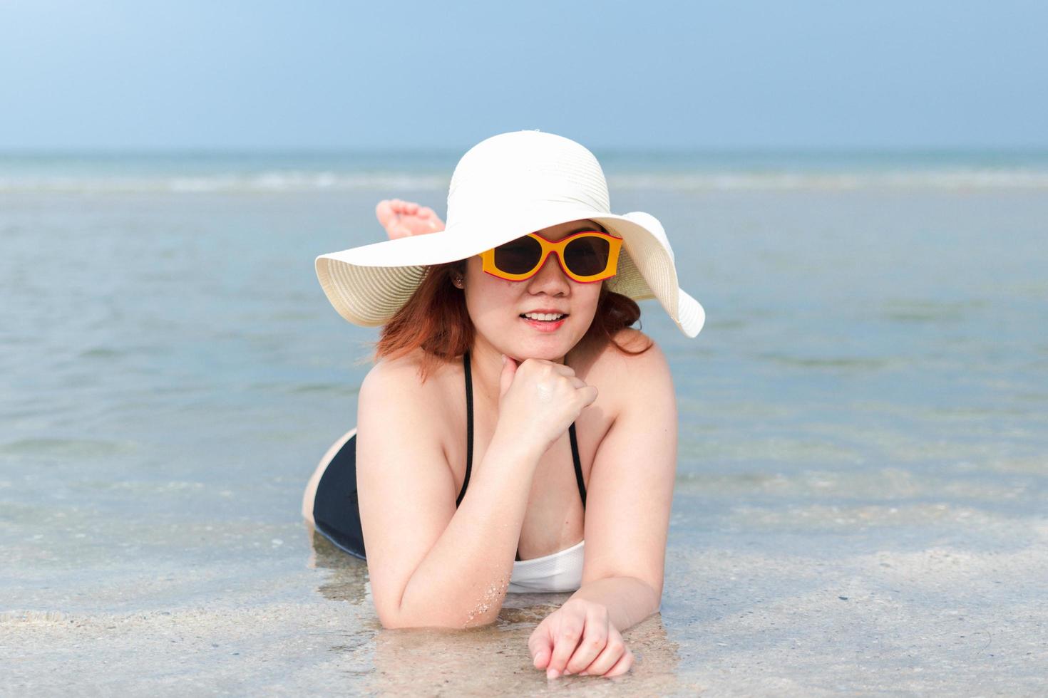 A plump white woman in a bathing suit, white hat, and yellow sunglasses is lying on the beach smiling prone. photo