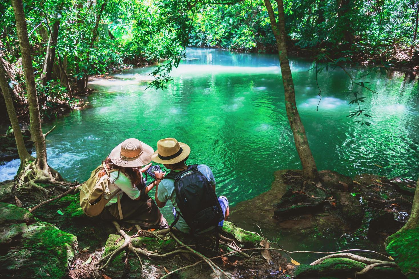 viajeros, parejas con mochilas, sentarse y relajarse en las rocas. viaja por la naturaleza en la jungla verde y disfruta de la vista en la cascada. turismo, senderismo, estudio de la naturaleza. parejas viajando tomando fotos