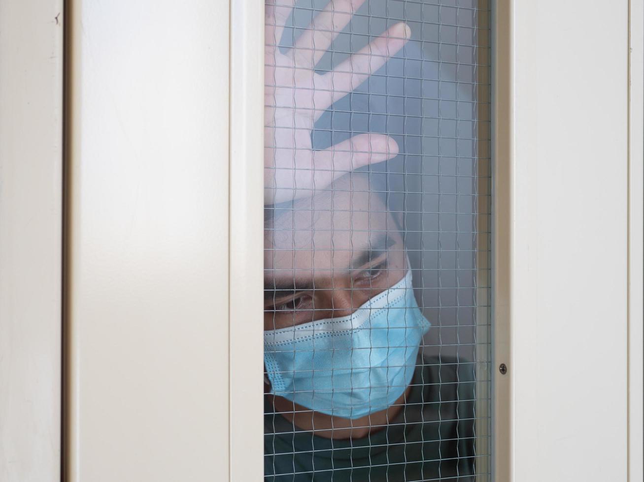 Lonely man in medical mask looking through the window. Isolation at home for self quarantine. Concept home quarantine, prevention COVID-19. Coronavirus outbreak situation photo