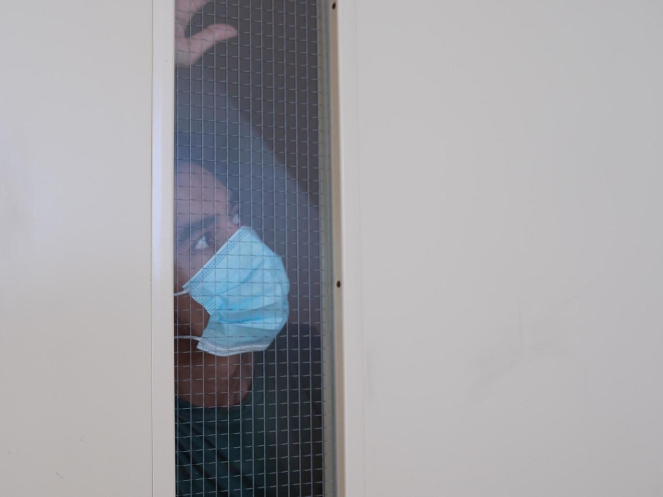 Lonely man in medical mask looking through the window. Isolation at home for self quarantine. Concept home quarantine, prevention COVID-19. Coronavirus outbreak situation photo