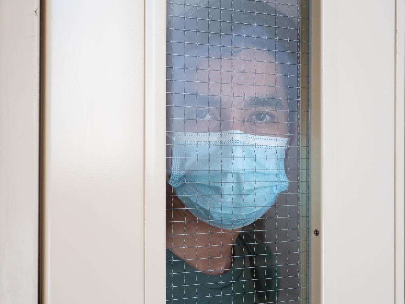 Lonely man in medical mask looking through the window. Isolation at home for self quarantine. Concept home quarantine, prevention COVID-19. Coronavirus outbreak situation photo