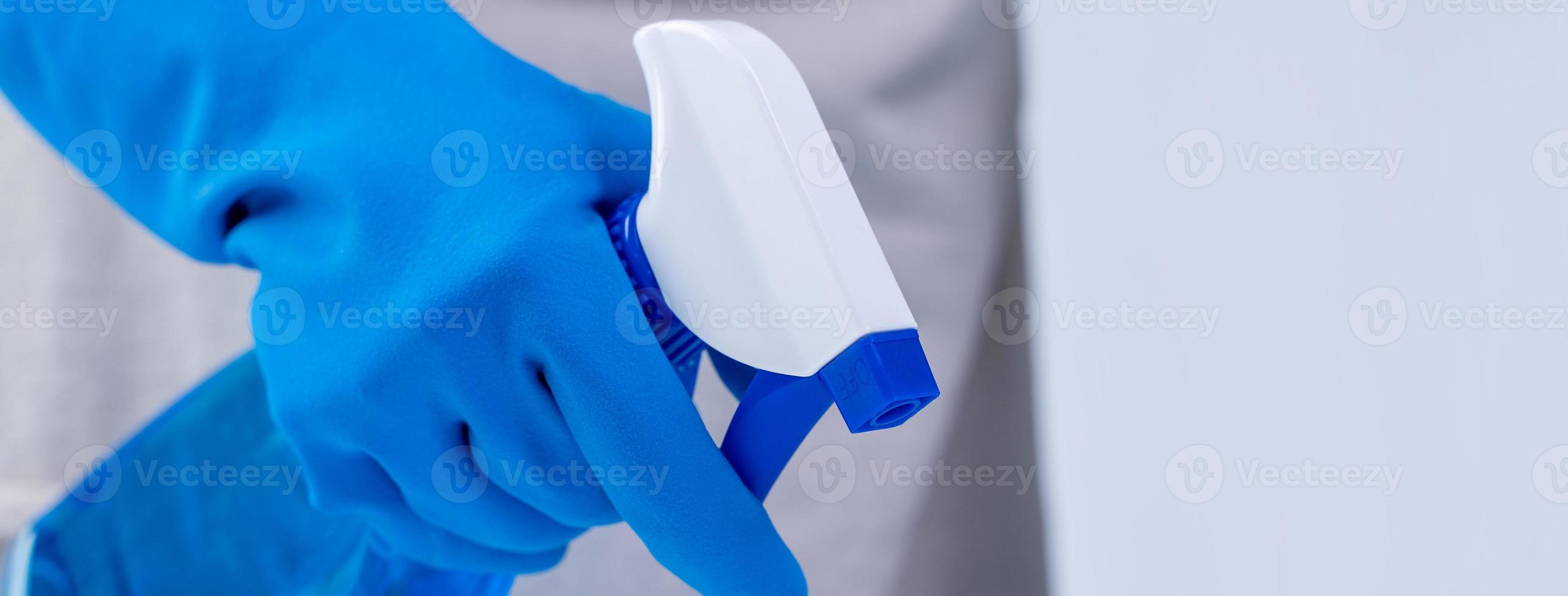 Young woman housekeeper in apron is cleaning, wiping down table surface with blue gloves, wet yellow rag, spraying bottle cleaner, closeup design concept. photo