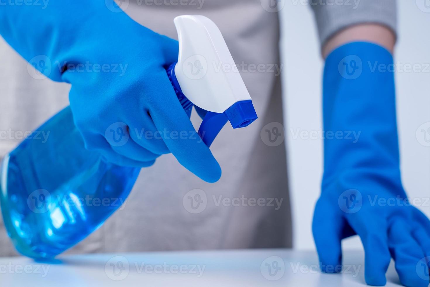 Young woman housekeeper in apron is cleaning, wiping down table surface with blue gloves, wet yellow rag, spraying bottle cleaner, closeup design concept. photo