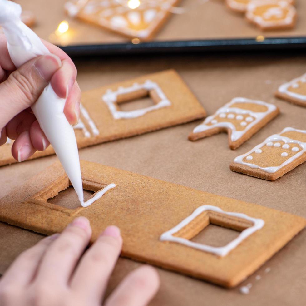 Young woman is decorating Christmas Gingerbread House cookies biscuit at home with frosting topping in icing bag, close up, lifestyle. photo