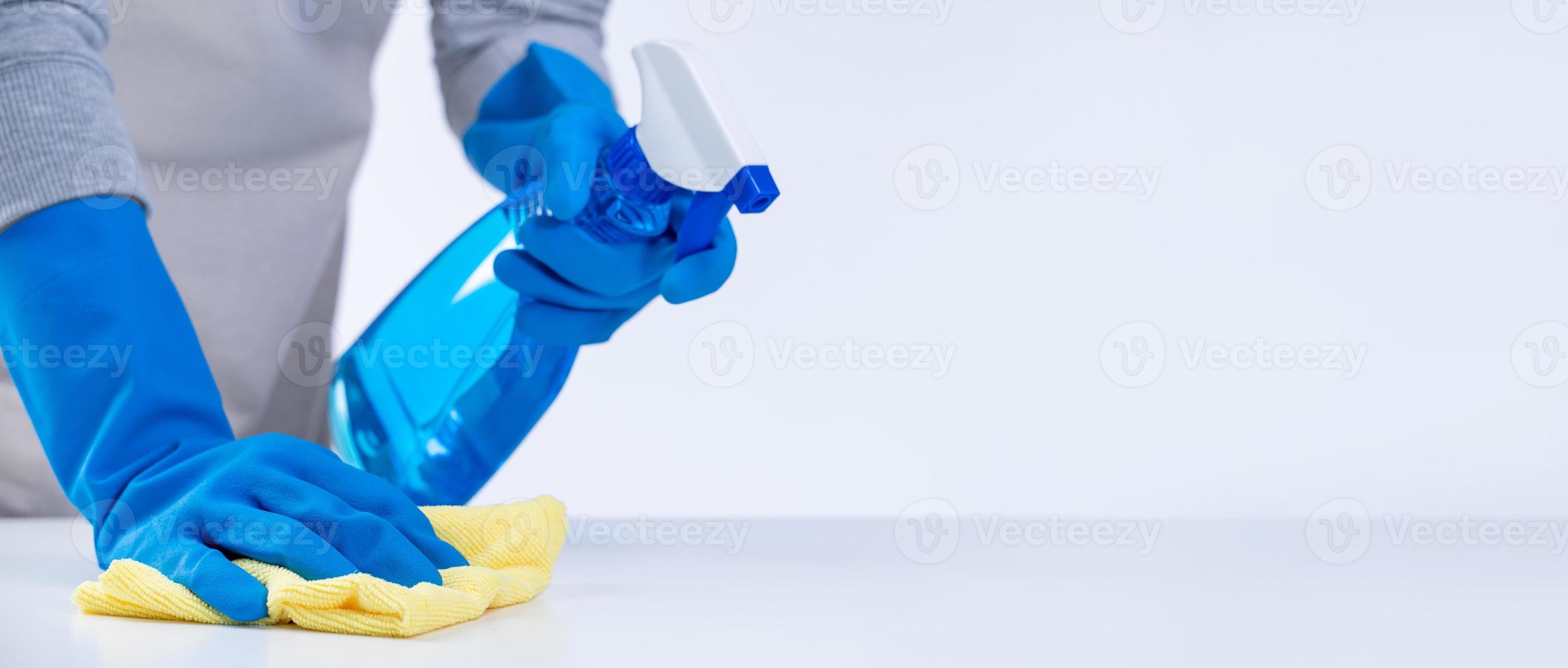 una joven ama de llaves en delantal está limpiando, limpiando la superficie de la mesa con guantes azules, trapo amarillo húmedo, limpiador de botellas rociadas, concepto de diseño cerrado. foto