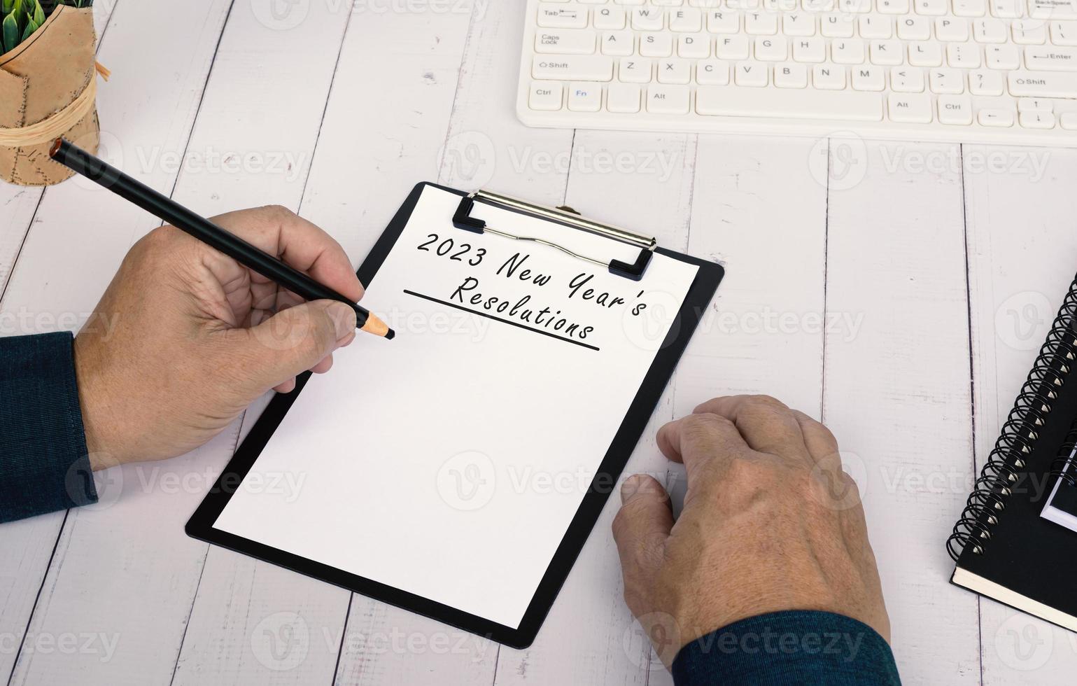 Hands writing a 2023 New Year's Resolutions list on a clip board on wooden desk. photo
