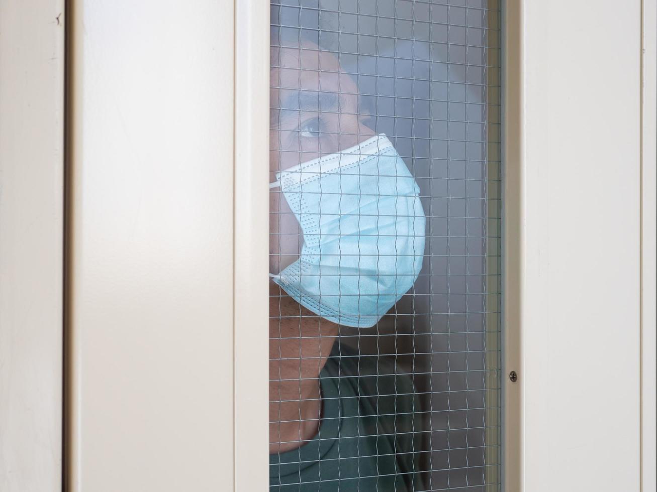 Lonely man in medical mask looking through the window. Isolation at home for self quarantine. Concept home quarantine, prevention COVID-19. Coronavirus outbreak situation photo