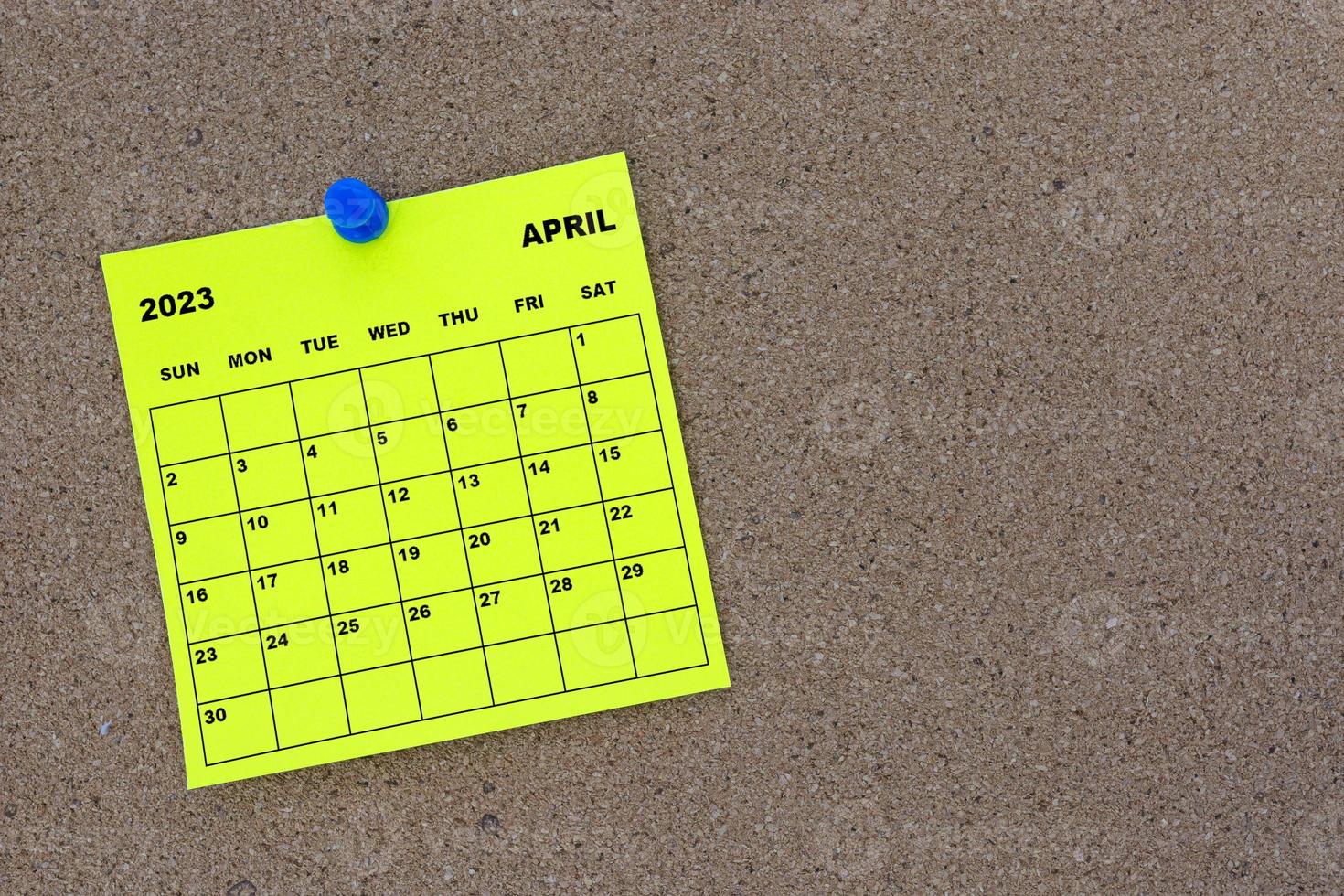 April 2023 yellow sticky note calendar with pin on cork bulletin billboard. photo