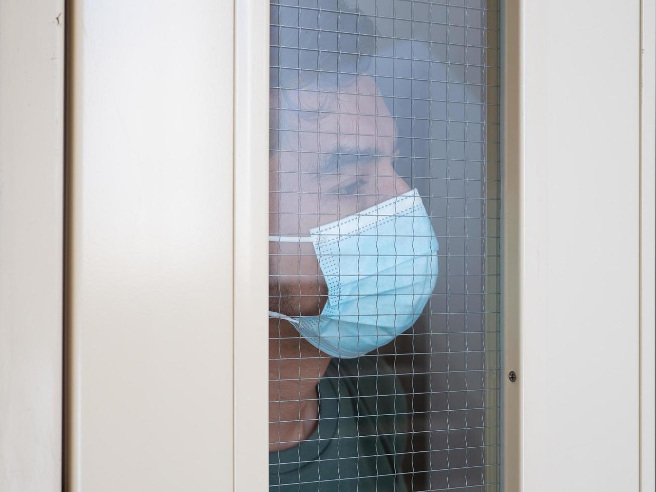 Lonely man in medical mask looking through the window. Isolation at home for self quarantine. Concept home quarantine, prevention COVID-19. Coronavirus outbreak situation photo