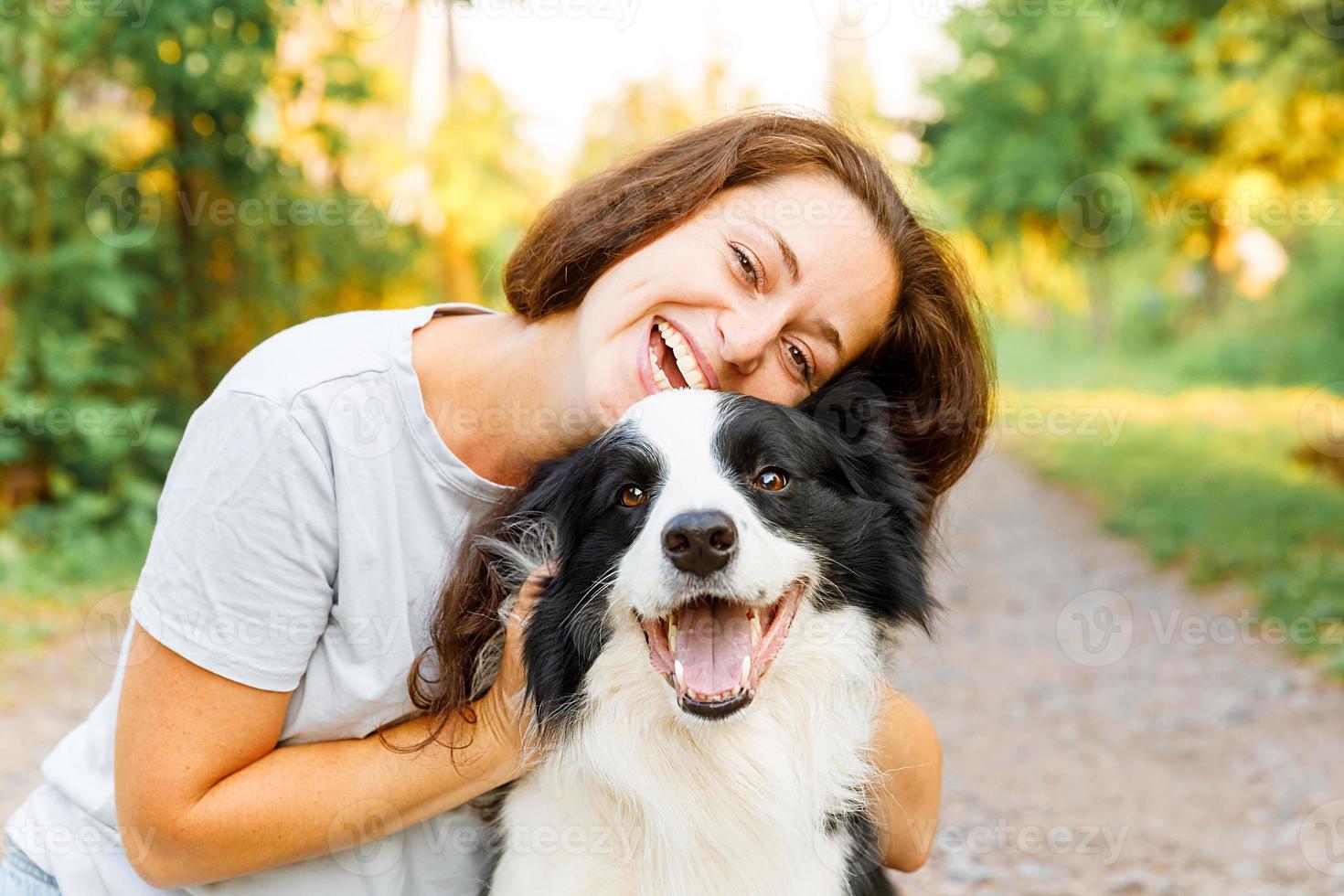 Smiling young attractive woman playing with cute puppy dog border collie on summer outdoor background. Girl holding embracing hugging dog friend. Pet care and animals concept. photo