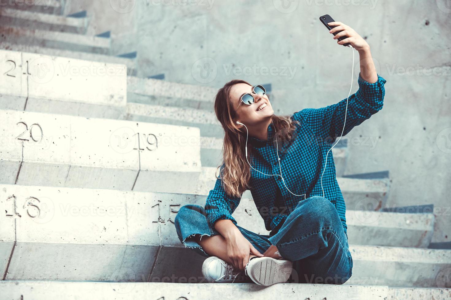 Photo of a cheerful smiling cute young student girl wearing sunglasses outdoors using mobile phone chatting listening music with earphones.