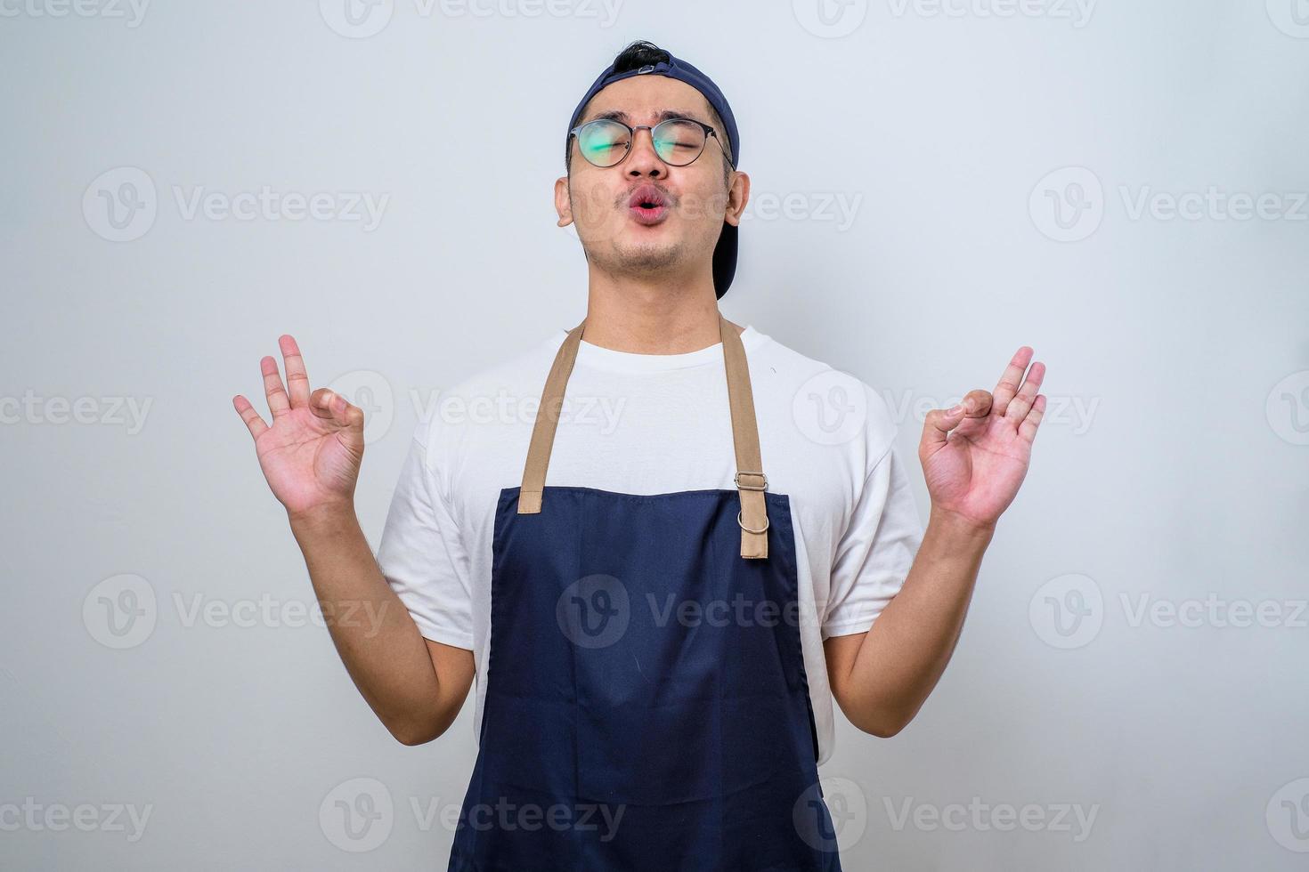 Young handsome asian barista man wearing apron standing relax and smiling with eyes closed doing meditation gesture with fingers. Yoga concept. photo