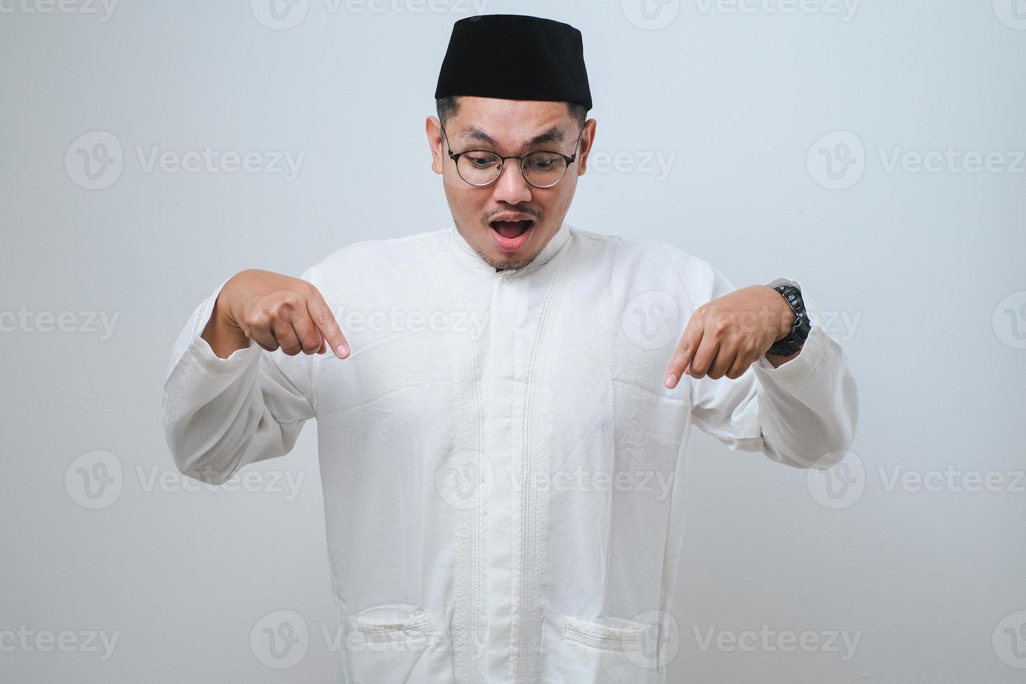 Asian muslim man feeling shocked, open-mouthed and amazed, looking and pointing downwards in disbelief and surprise against white background photo