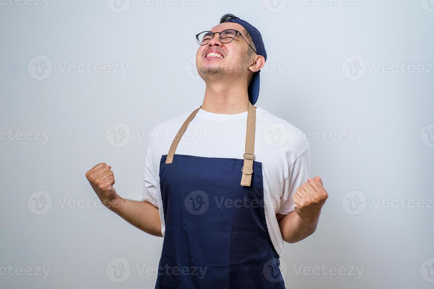 Asian barista man wearing apron standing very happy and excited doing winner gesture with arms raised, smiling and screaming for success. Celebration concept. photo