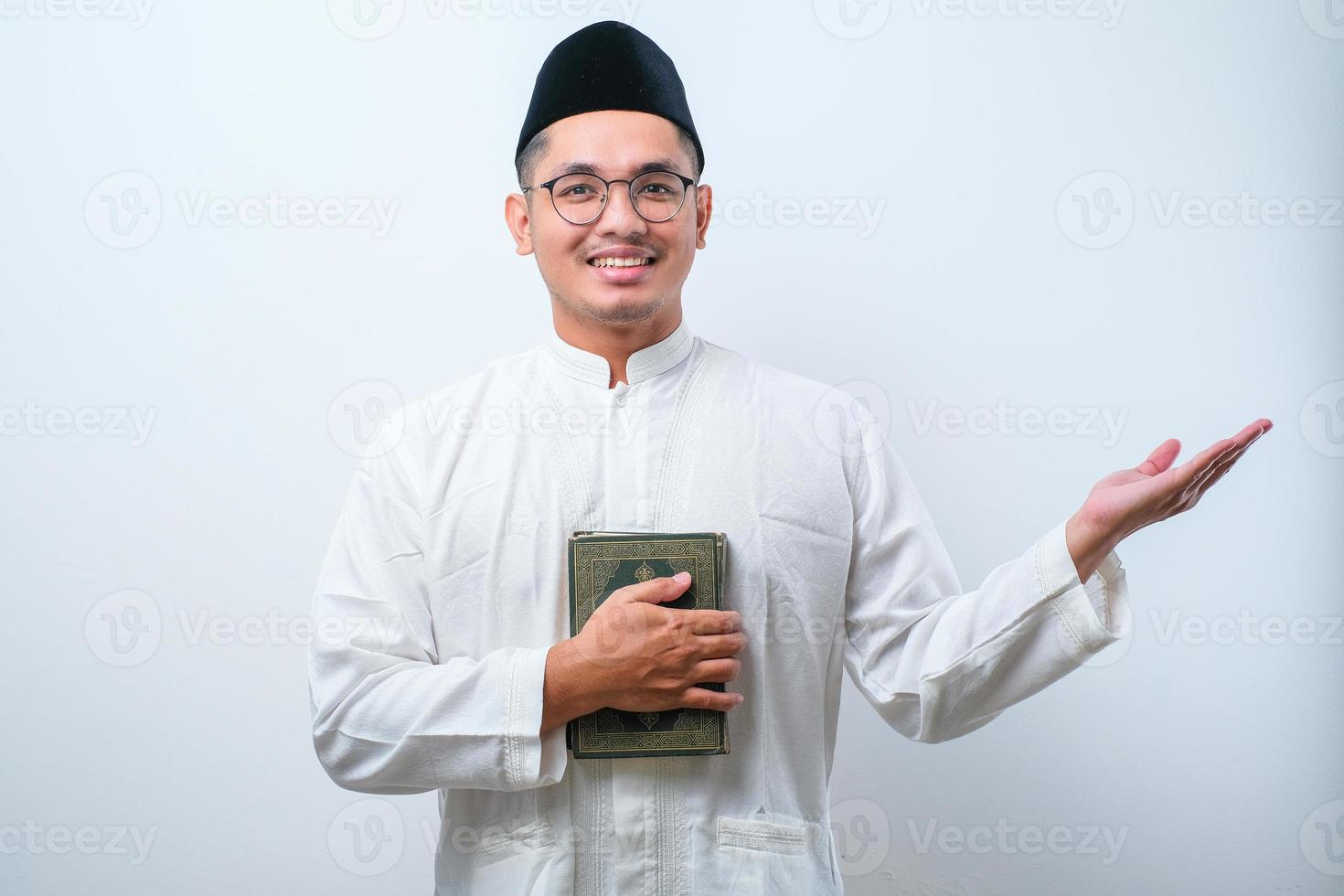 Asian muslim man smiling holding al quran book in hands and showing something on his side photo