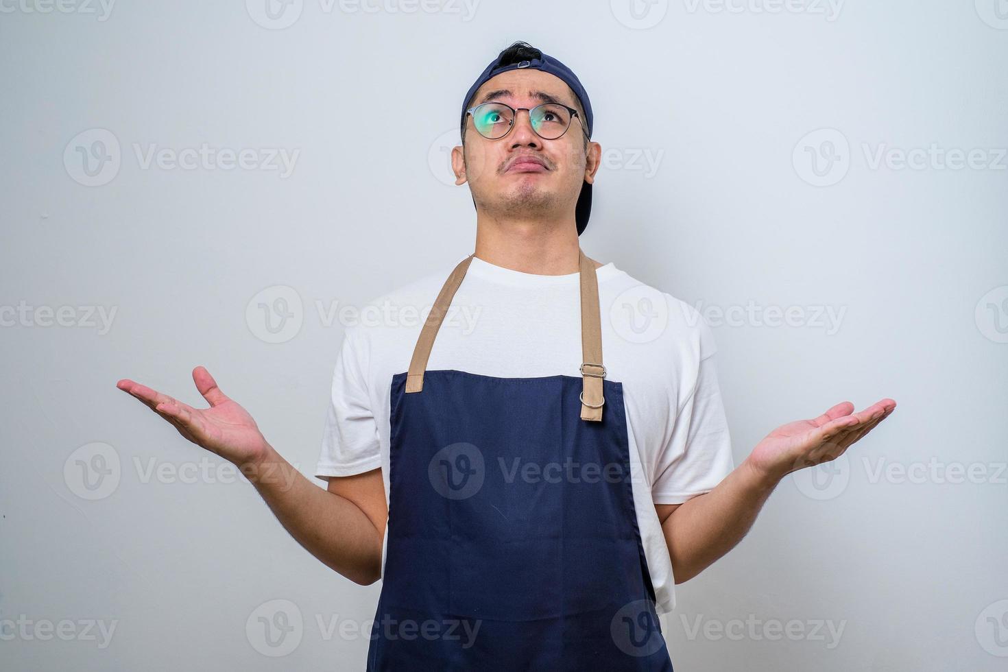 Young asian barista man wearing apron clueless and confused expression with arms and hands raised photo