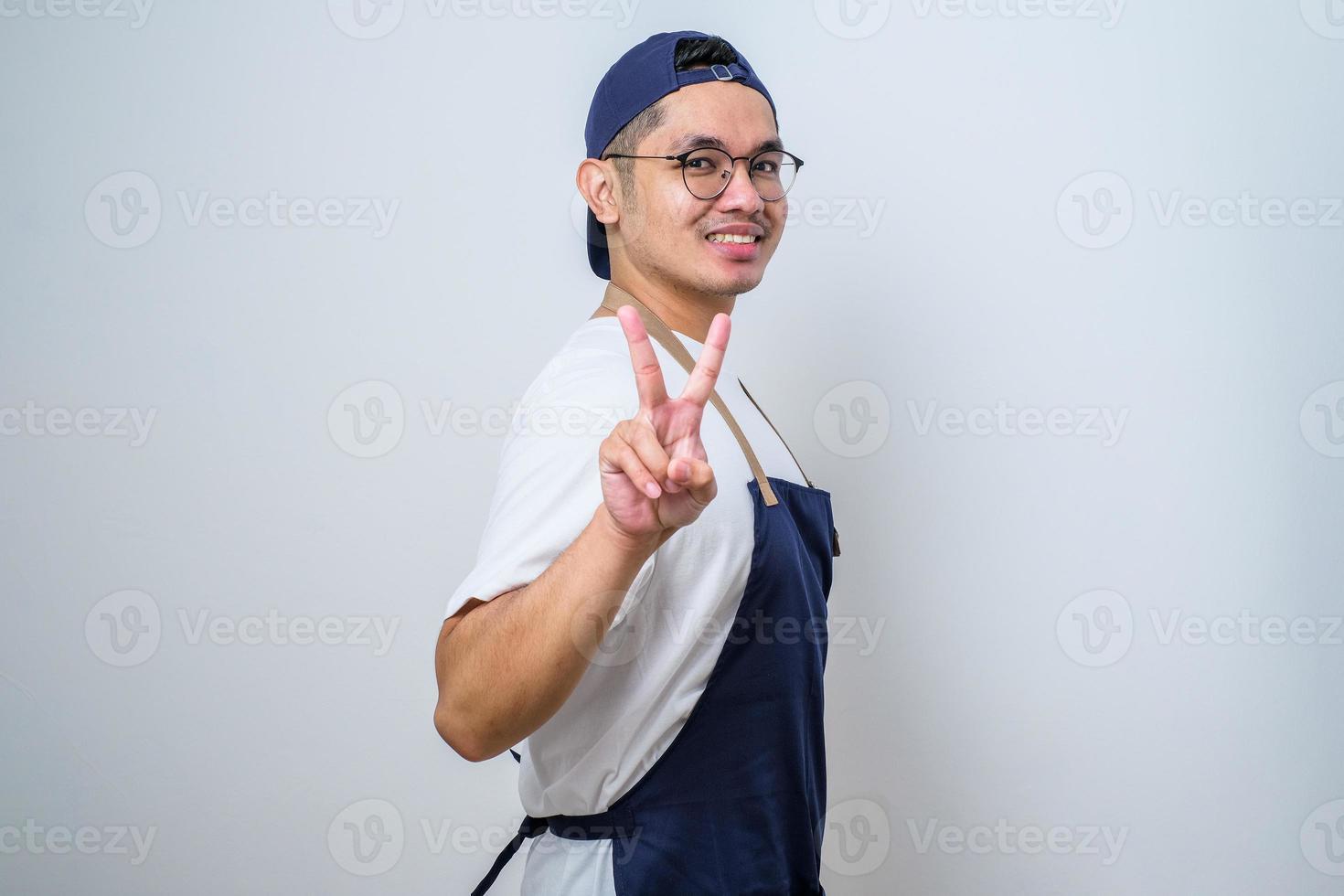 Young asian barista man wearing glasses and cap doing peace symbol with fingers over face, smiling cheerful showing victory photo