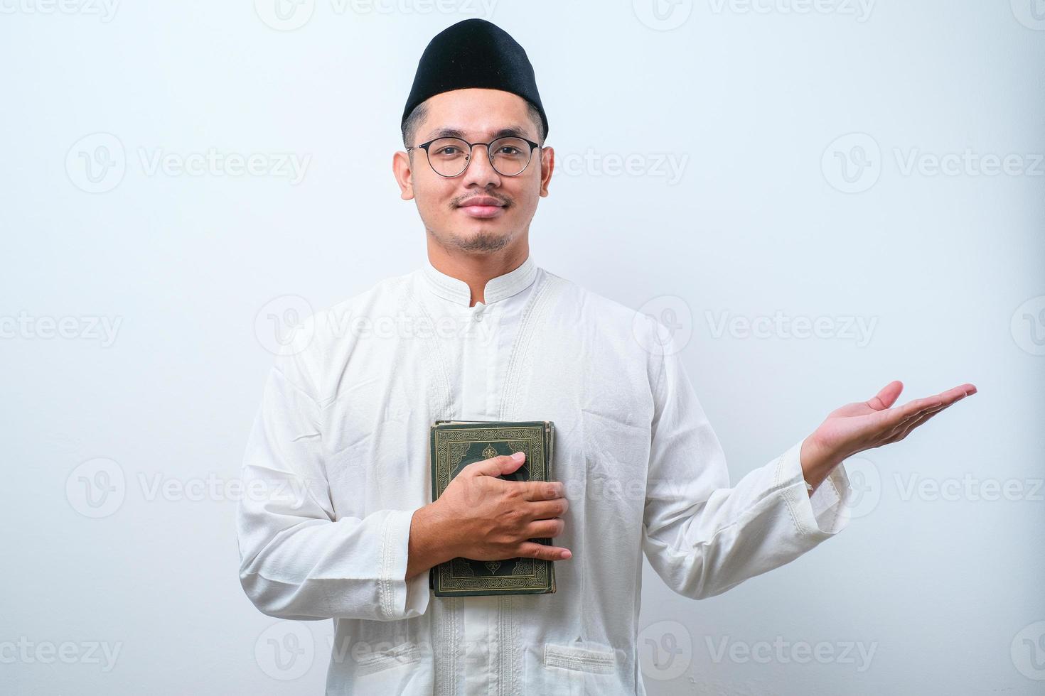Asian muslim man smiling holding al quran book in hands and showing something on his side photo
