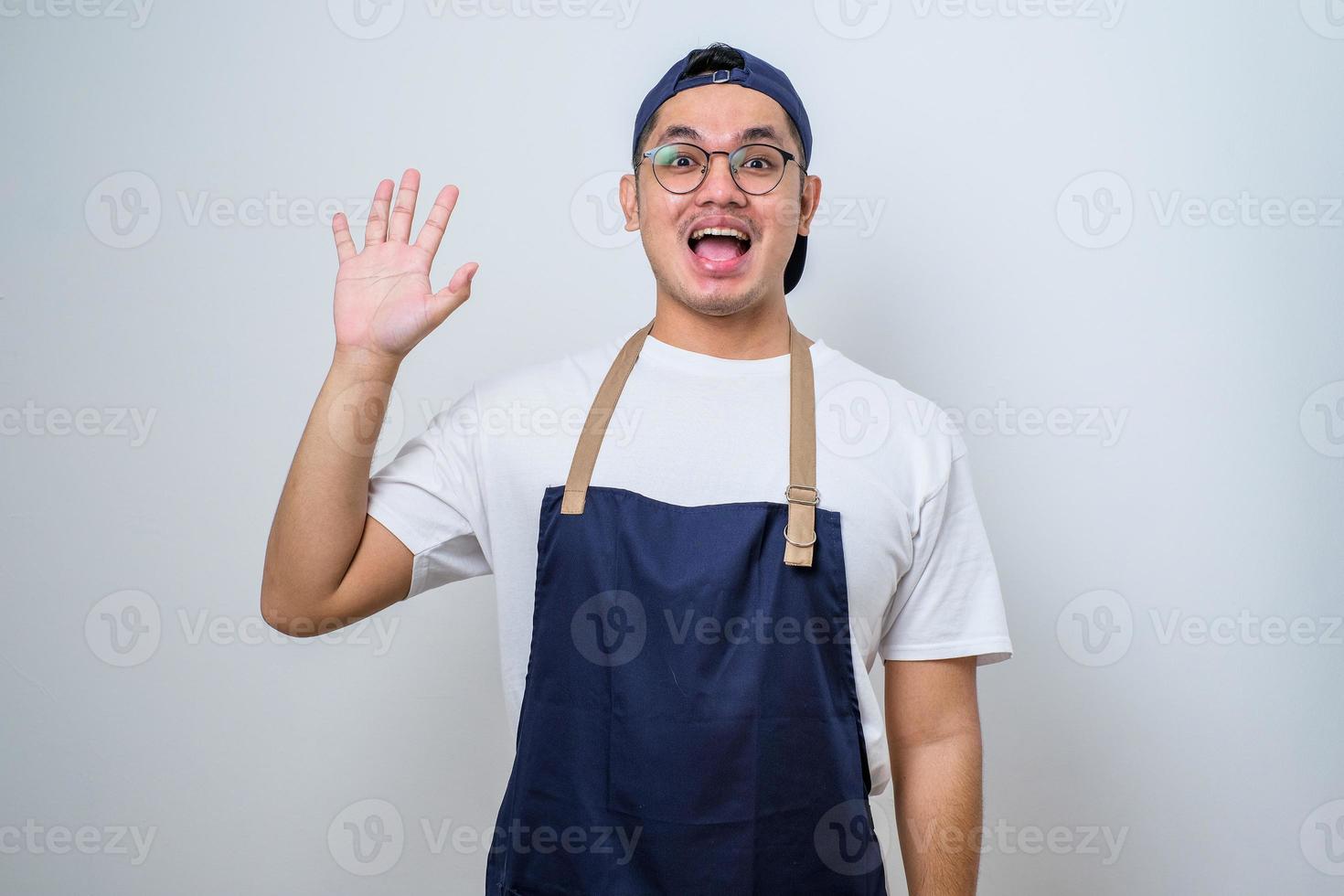 Young asian barista man wearing apron greeting client waving to say hello or hi to customer photo