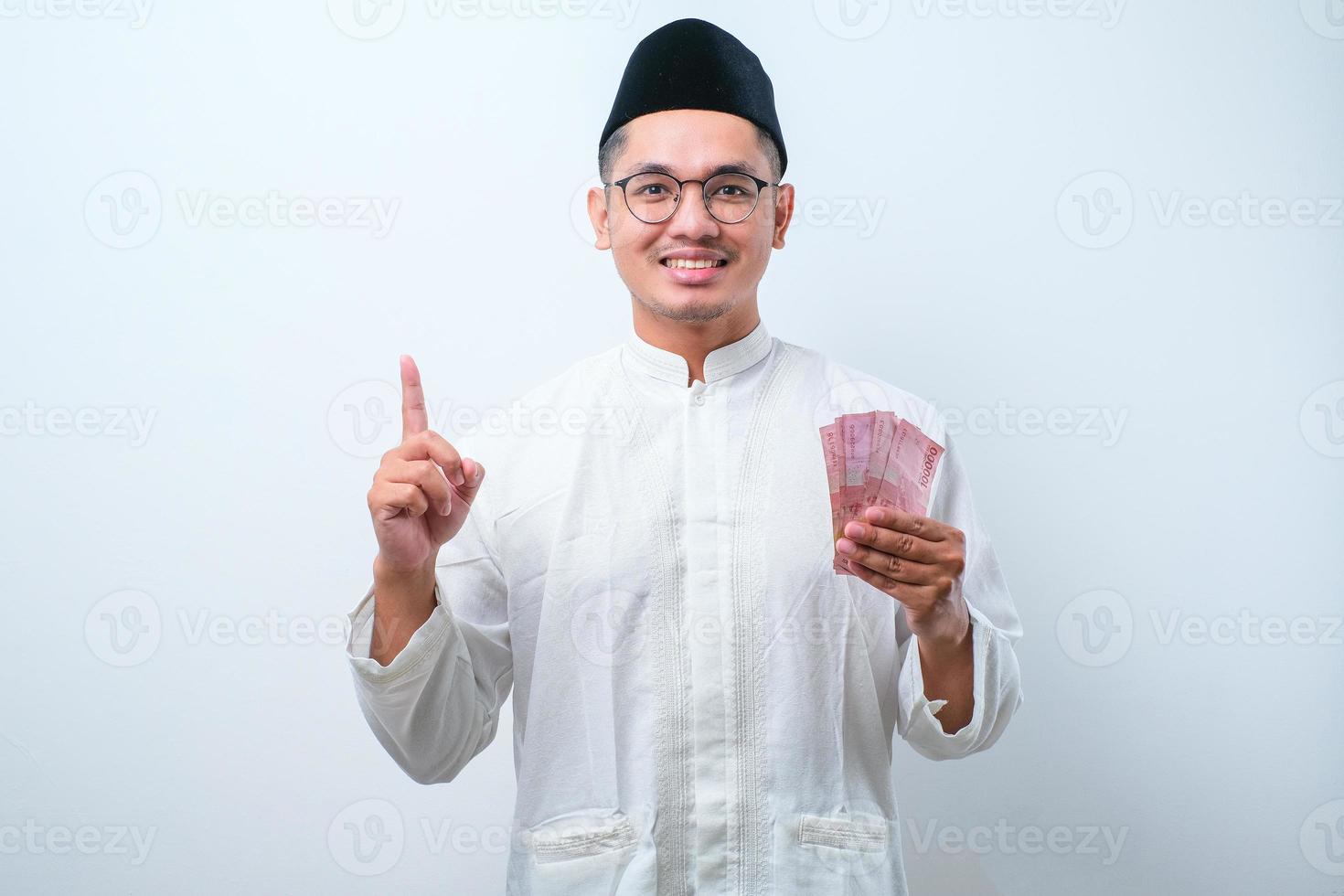 Asian muslim man holding rupiah paper money while pointing his finger up and smiling photo