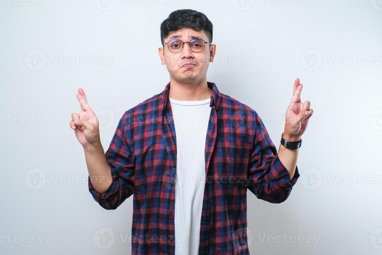 Portrait of a young handsome Asian man wearing casual shirt and glasses doing fingers crossed, wishing and praying for miracle. photo