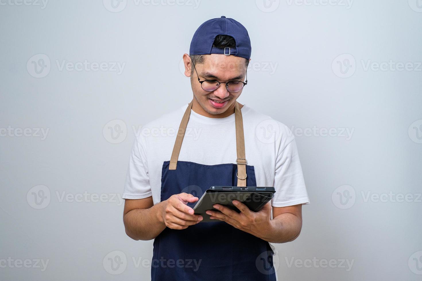 Cheerful young Asian barista man wearing apron and cap using digital tablet to manage cafe order photo