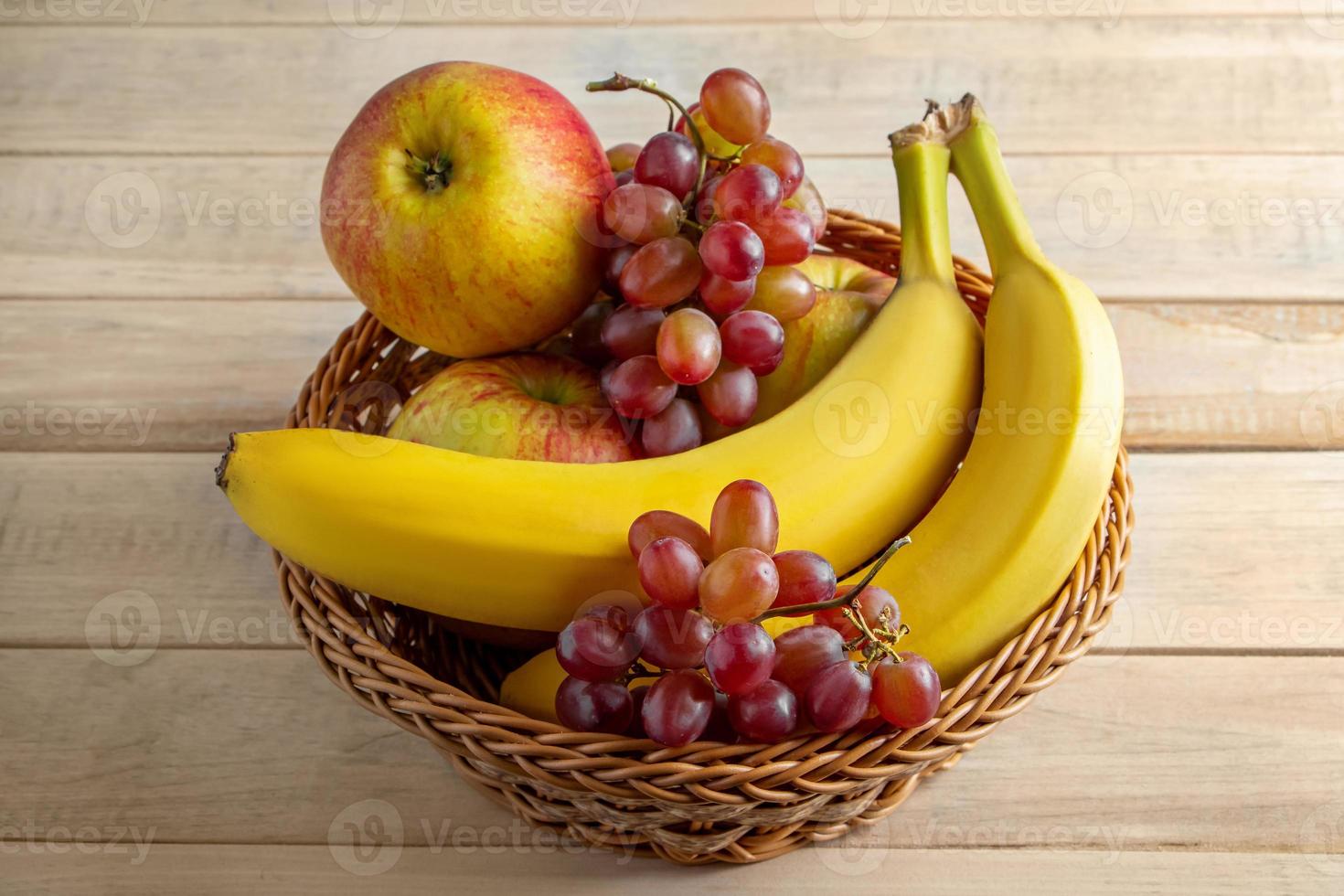 Ripe fruits in wicker basket on wooden background. Bananas, apples and grapes. Harvest concept. photo