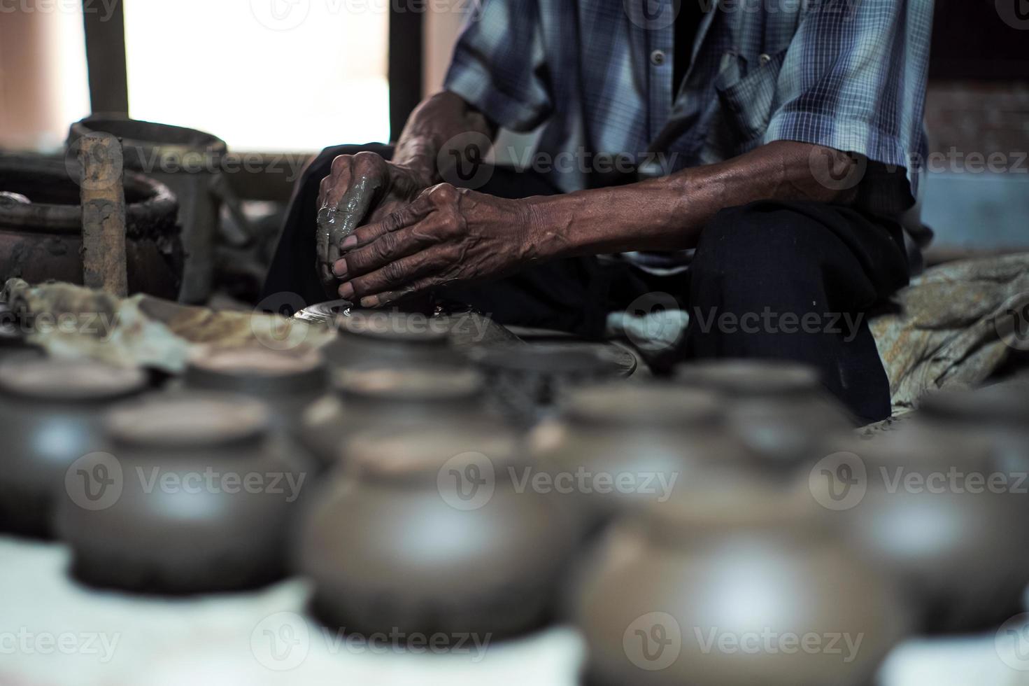 Selective focused on the dirty wrinkled skin hands of old man molding the clay work on the spinning wheel for making the jar with blurred group of clay jars in foreground photo