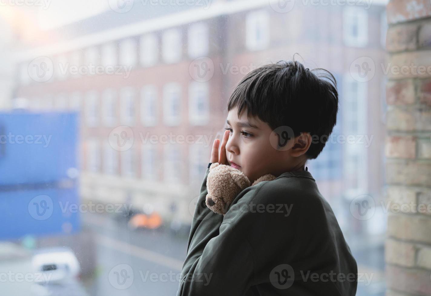 Lonely Kid with sad face hugging teddy bear standing next to window, Bored Child holding soft toy standing alone looking through window glass, Young boy looking out deep in thought. photo