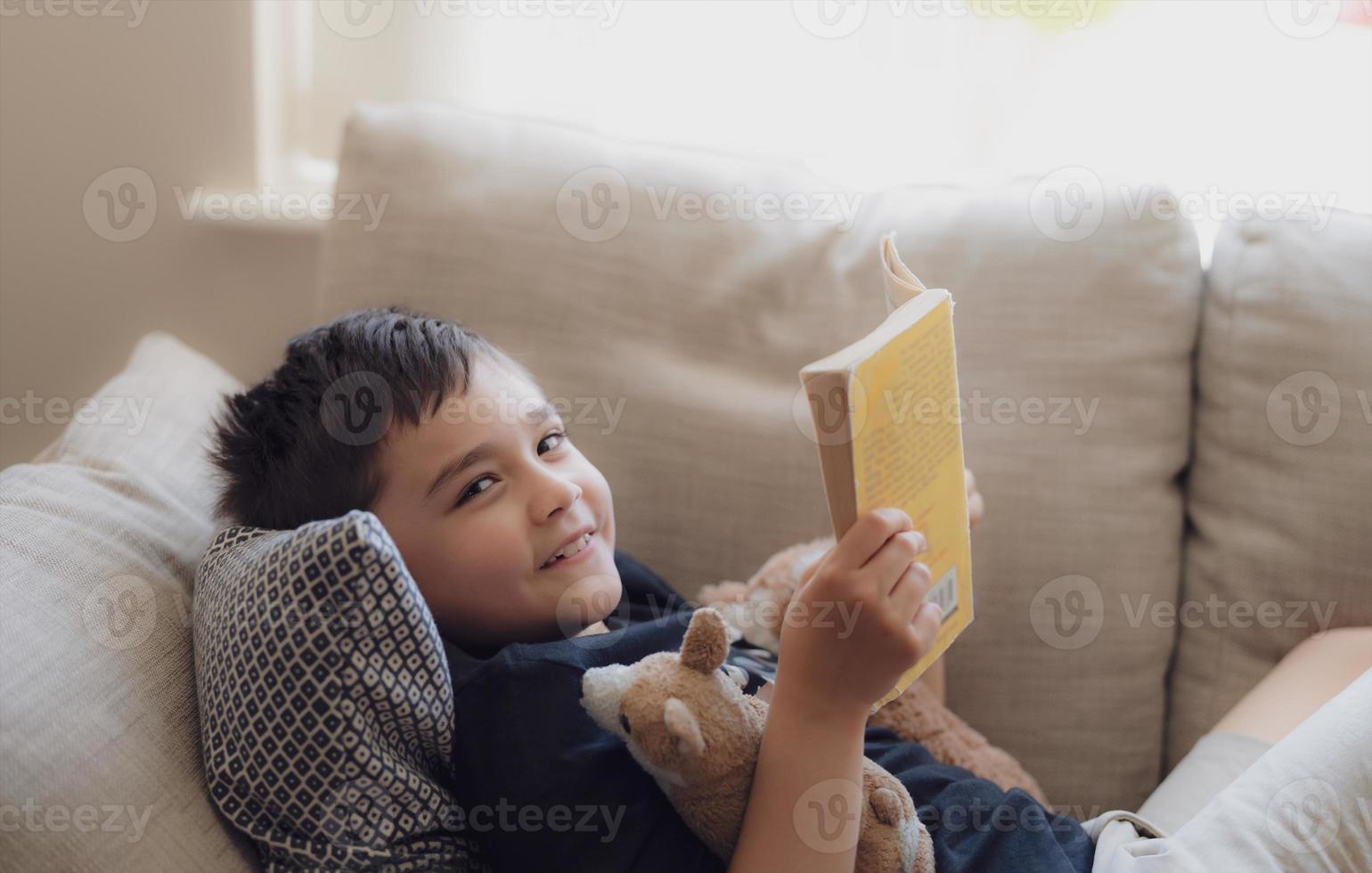 niño de la escuela de retratos leyendo un libro para la tarea, niño acostado en el sofá disfruta leyendo la historia en la sala de estar, niño relajándose en casa en la primavera soleada, educación en el hogar o concepto de educación foto